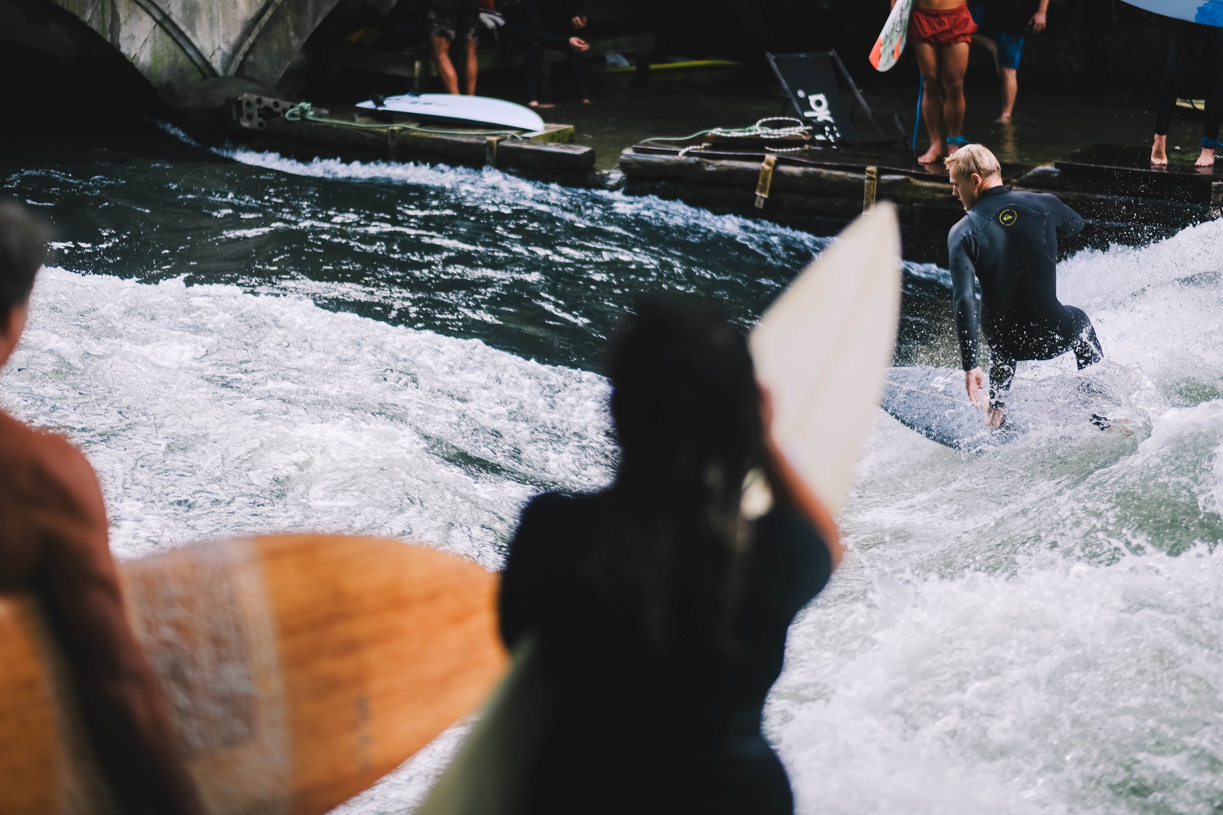 Surfers preparing to ride a wave in a river, with a person surfing in the foreground and others holding surfboards on a platform nearby.