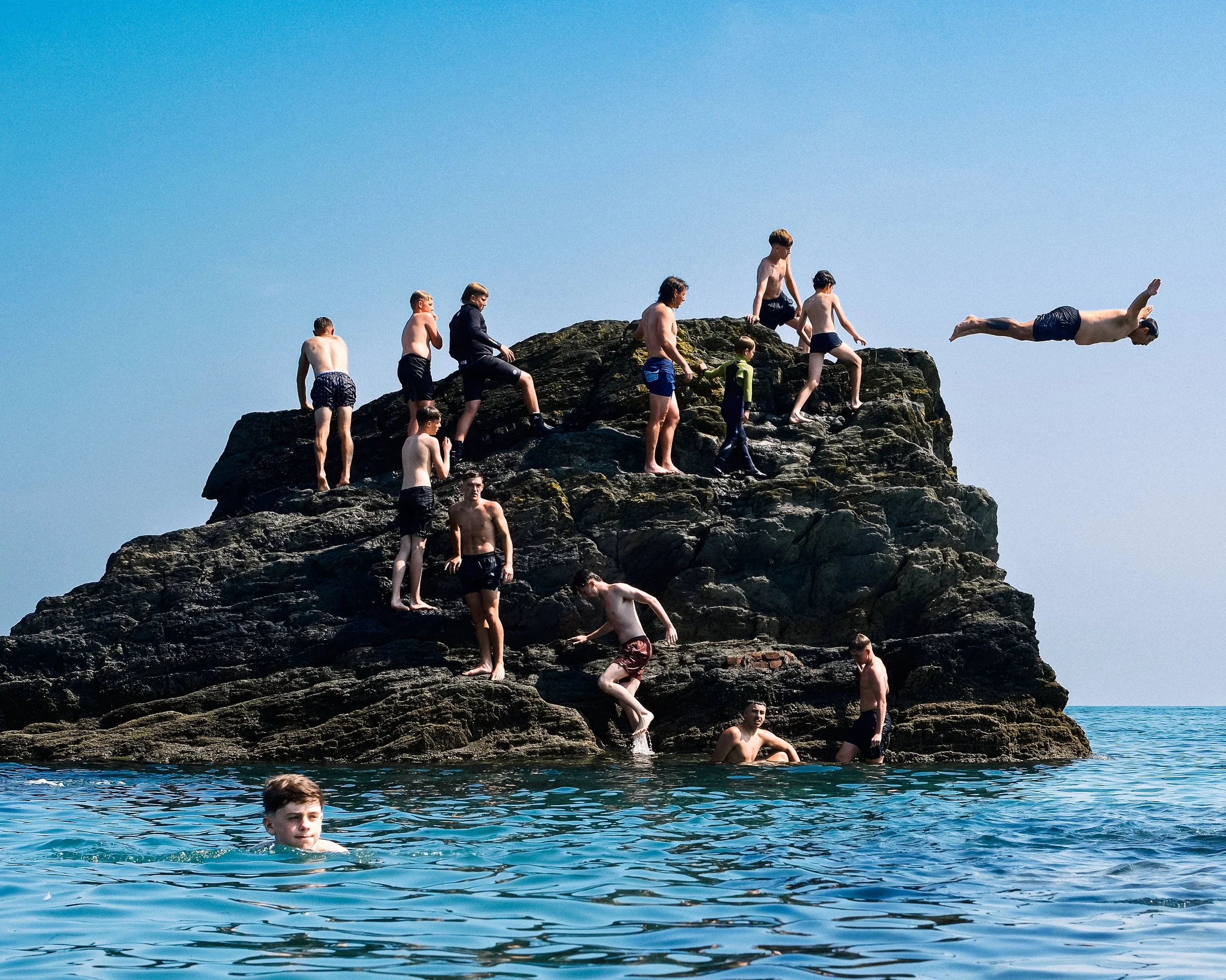 People jumping off and climbing on a rocky formation in the water on a sunny day.