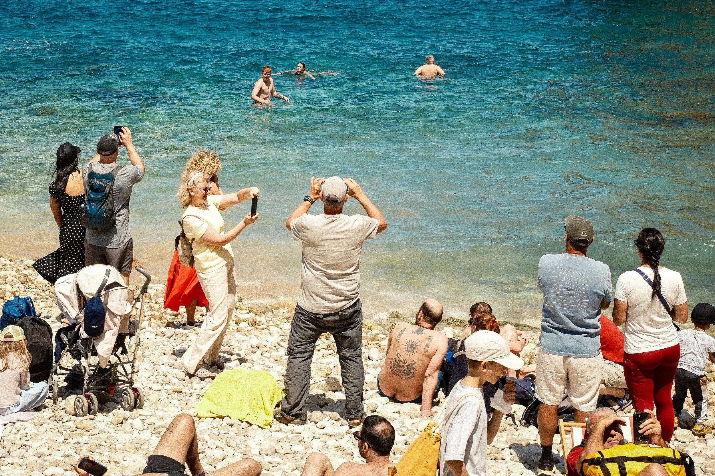People gathered on a rocky beach, some taking photos or videos, while others are swimming or relaxing in the water.