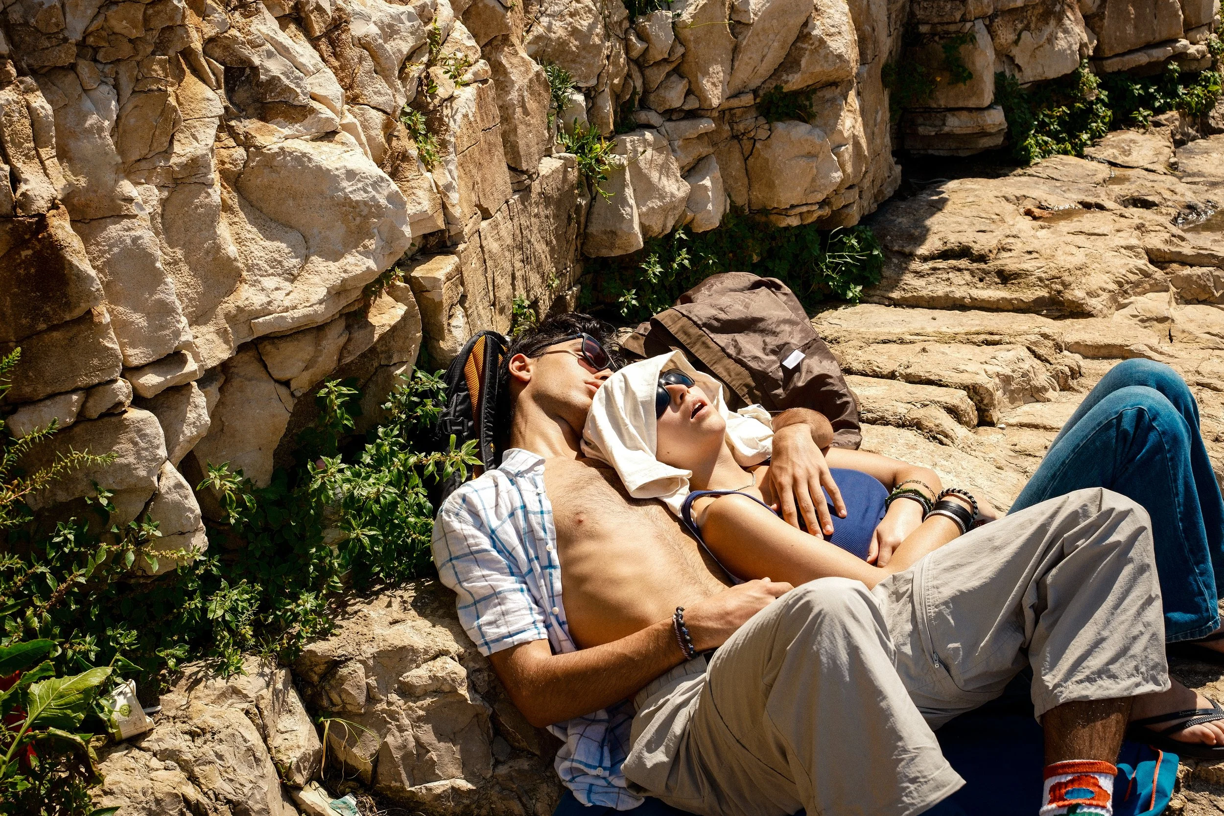 Two young people lie on the ground resting against a stone wall. They are wearing sunglasses and casual summer clothing, with a backpack nearby. The scene appears to be outdoors on a sunny day.