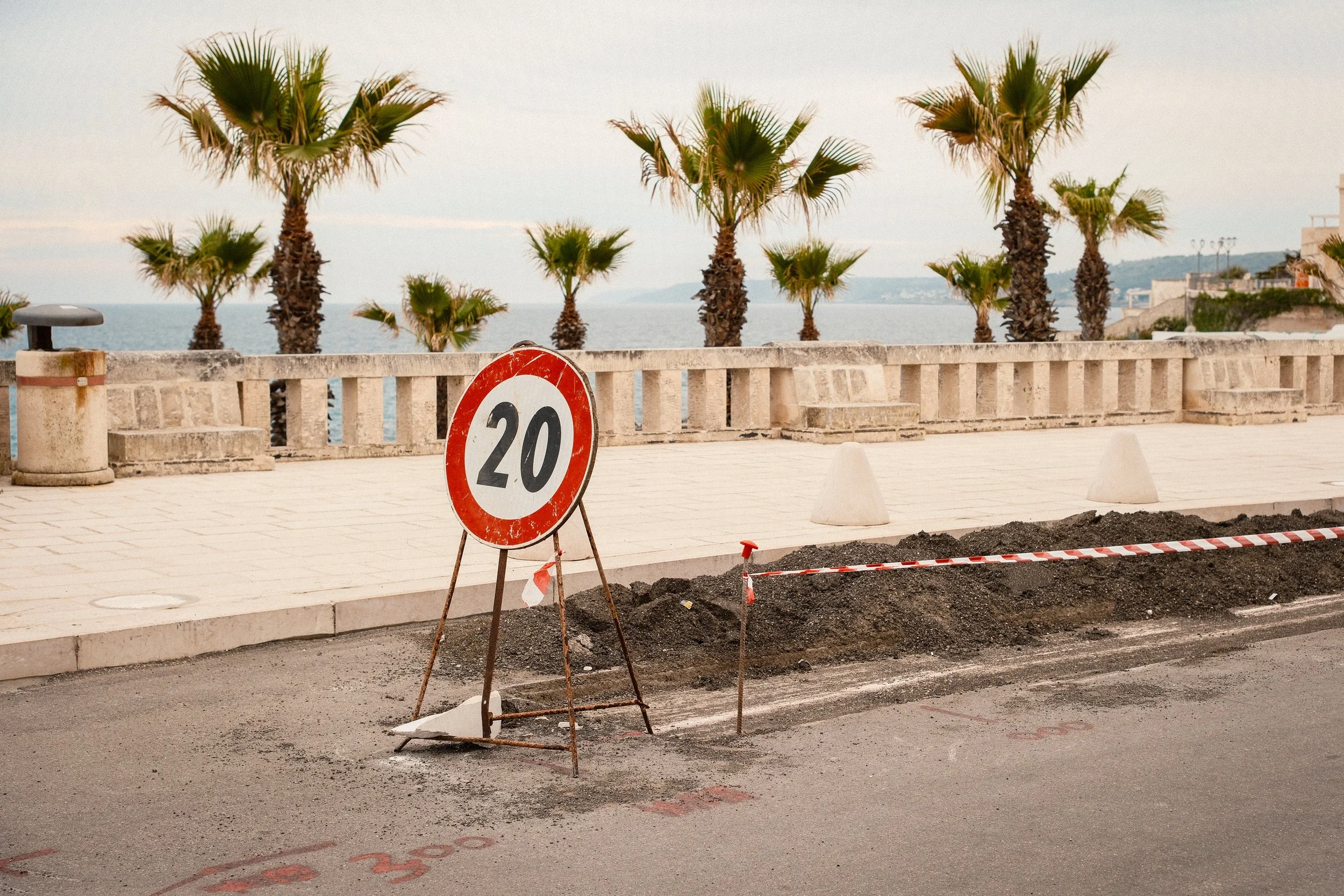 Construction area with a red and white 20 mph speed limit sign, a concrete wall, and palm trees in the background by the sea.