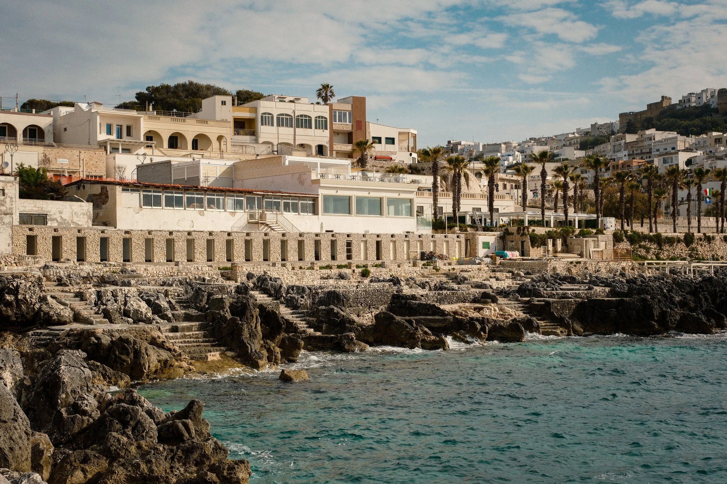 Seaside view showing rocky shore in the foreground, with buildings and palm trees on a hillside in the background, under a partly cloudy sky.