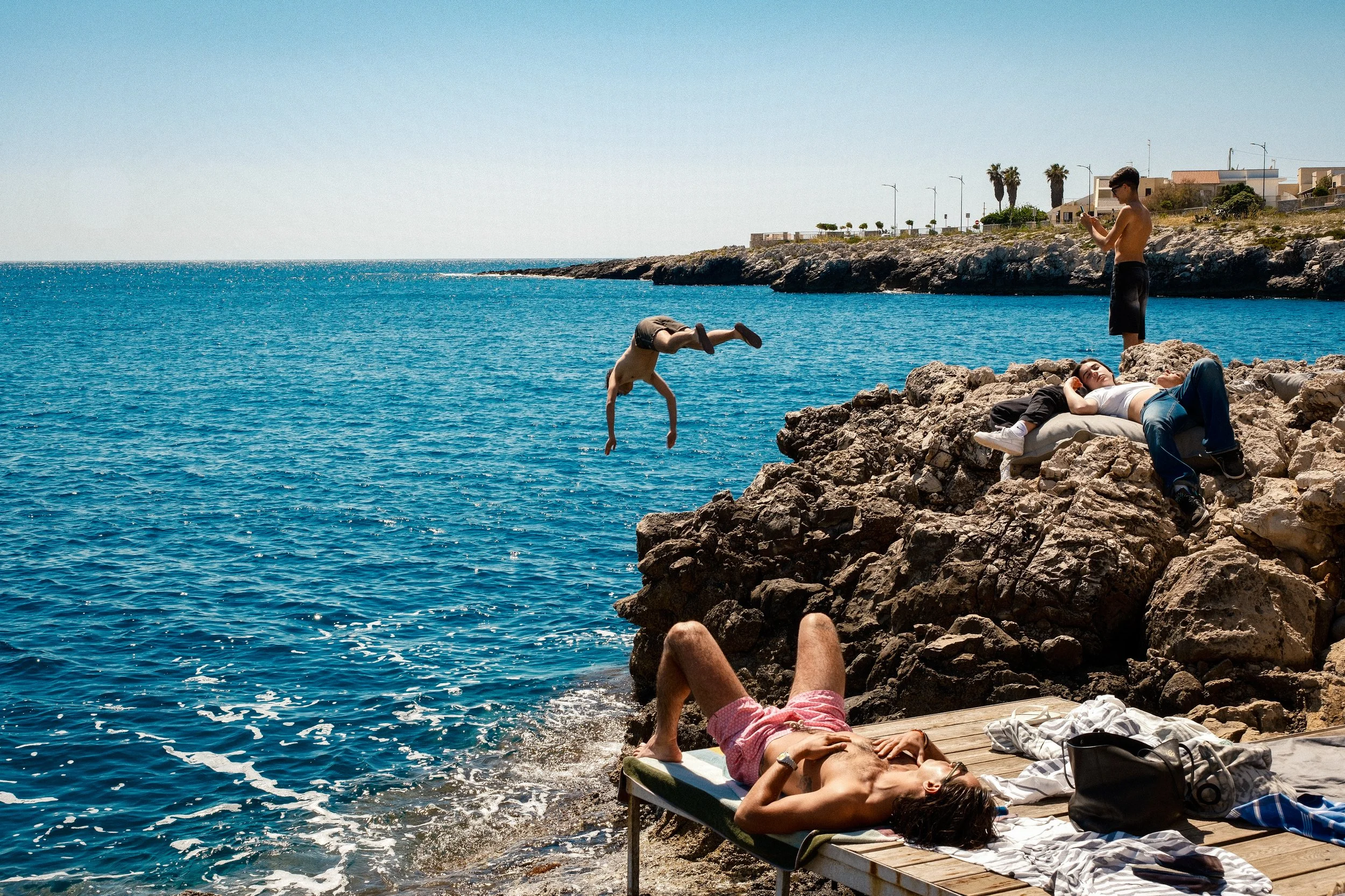 People relaxing and playing on rocky coast near blue ocean, with one person performing a handstand, others laying on rocks and a bed, someone standing and using a phone, in a sunny coastal area with houses and palm trees in the background.