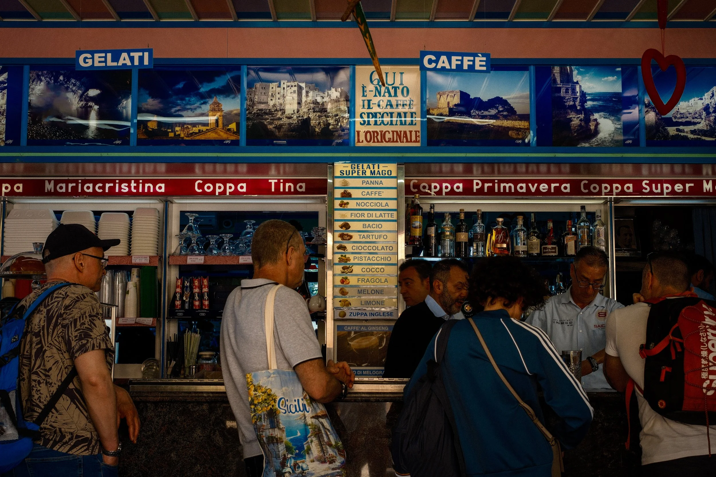 A crowded Italian food and drink stall with signs advertising gelato and coffee, several people standing in line, and scenic travel posters in the background.