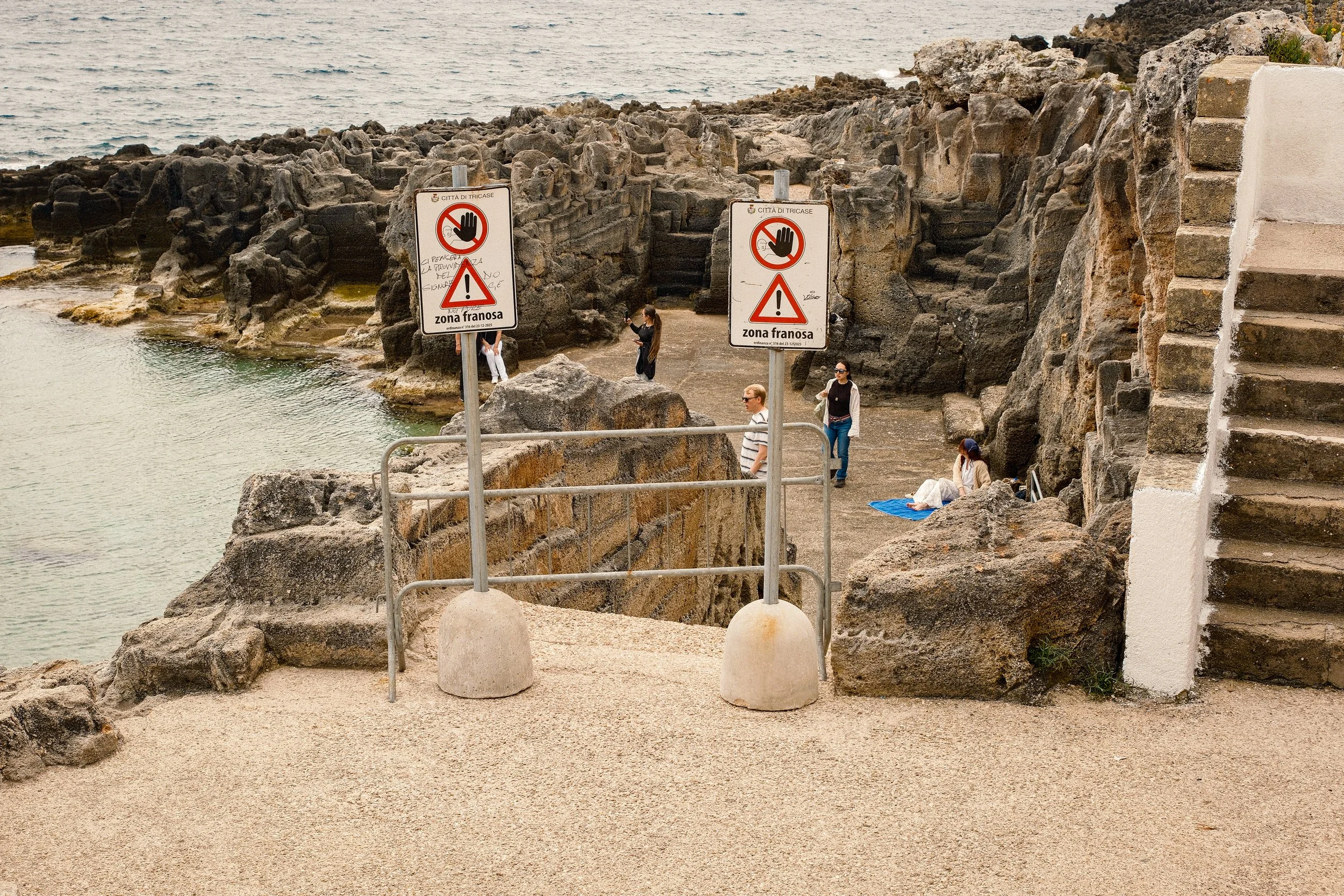 People by the rocky coast with two warning signs in Italian, indicating a dangerous zone and no entry. Some individuals are sitting and walking near the water, with steps leading down to the shoreline, and the ocean in the background.