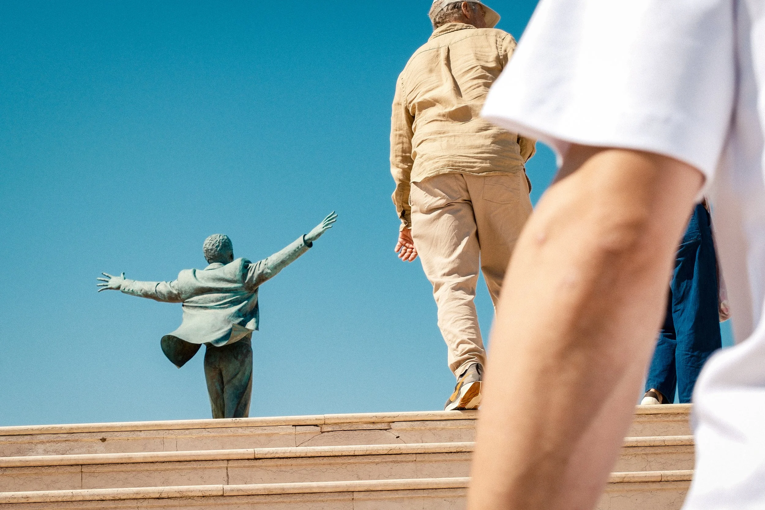 People walking up stairs towards the statue of a man with outstretched arms against a blue sky.