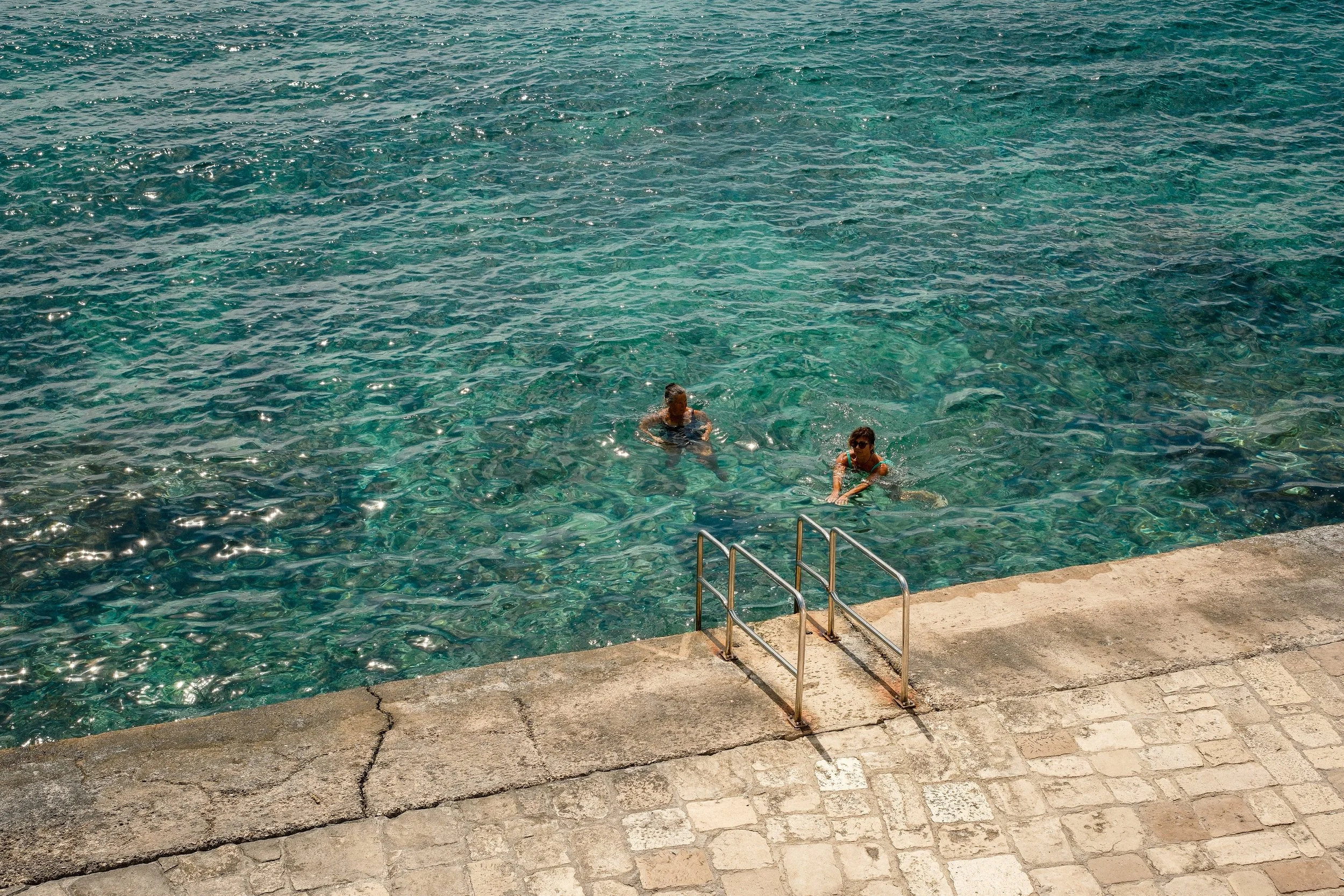 Two people swimming in a clear, blue water swimming pool with a concrete deck and metal handrails.