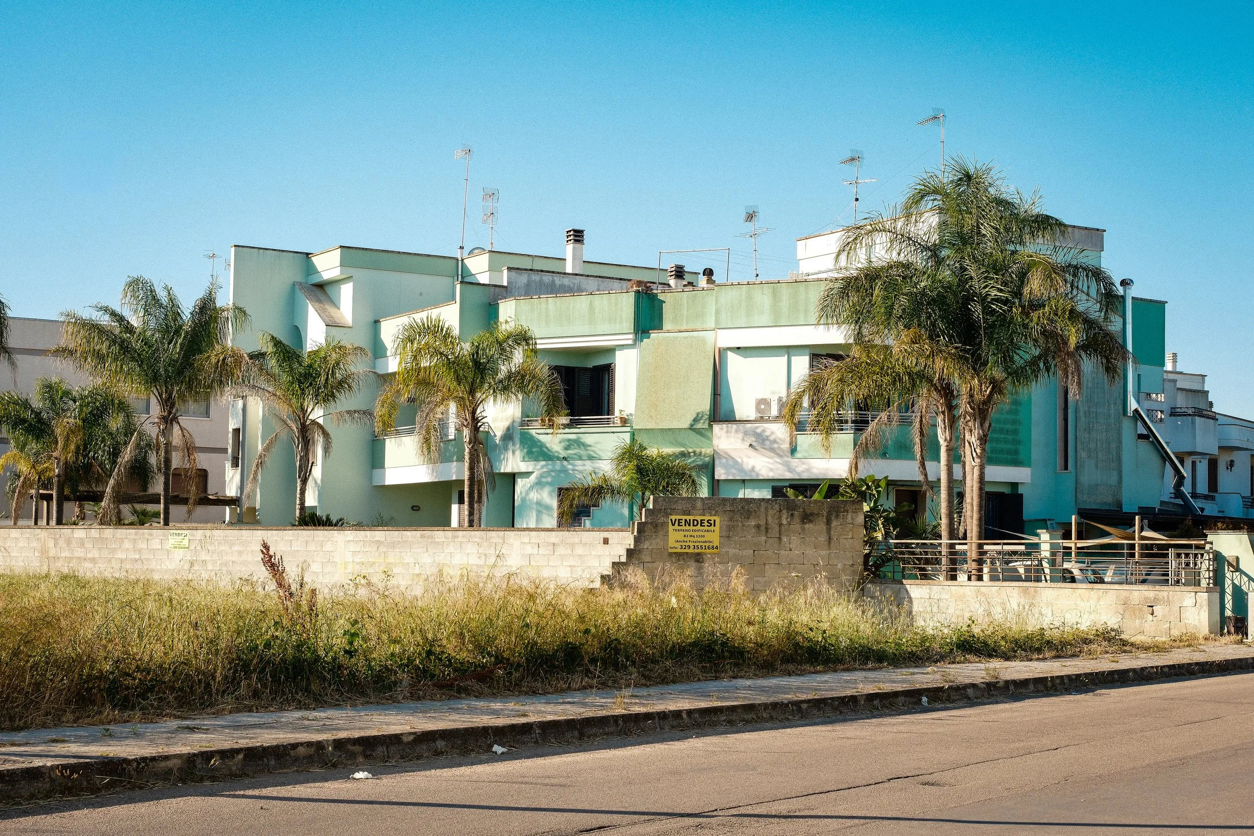 A multi-story pastel green and white apartment building with palm trees in front, under a clear blue sky, and a concrete wall with a "VENDESI" sign indicating it is for sale.