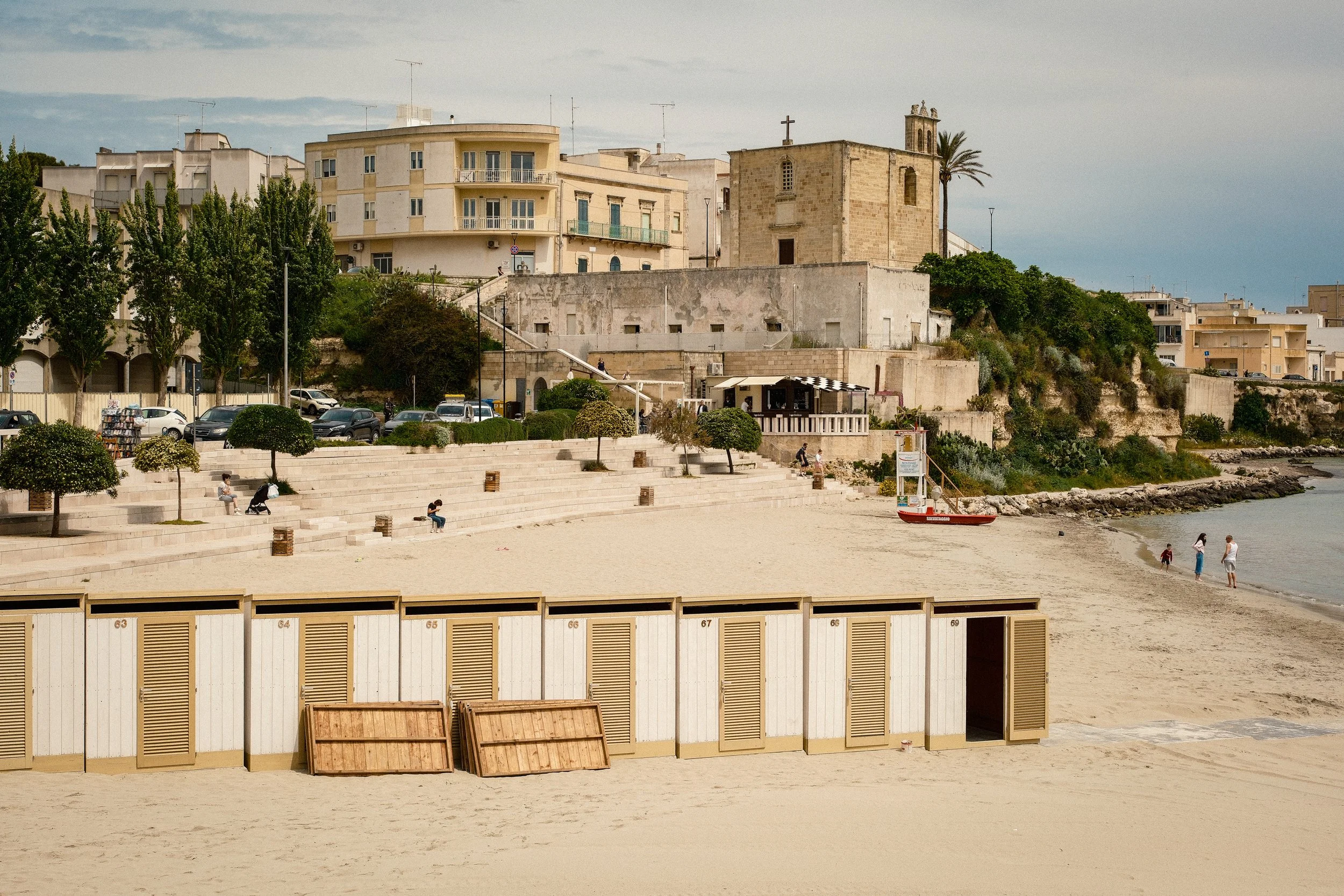 Beach with row of beach huts, some are open and others closed, along the sandy shore. Several people are walking or sitting, and there is a small boat near the water. In the background, there are multi-story buildings and an old stone church on a hill with trees around.