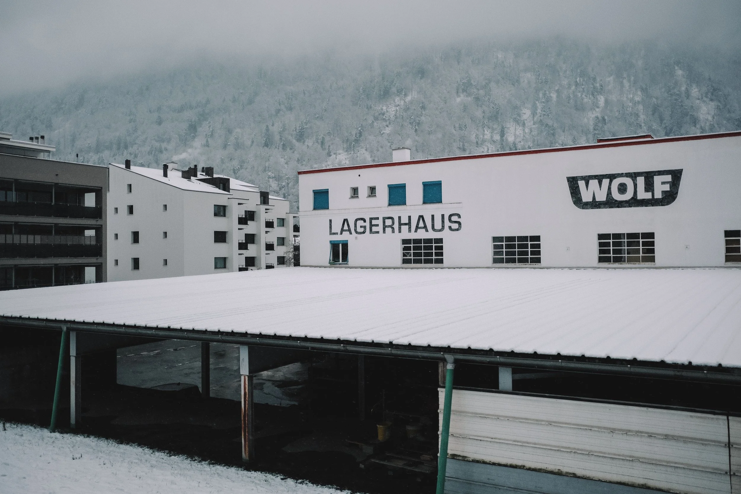 Snow-covered warehouse building with 'LAGERHAUS' and 'WOLF' signage, surrounded by residential buildings and snowy mountains in the background.