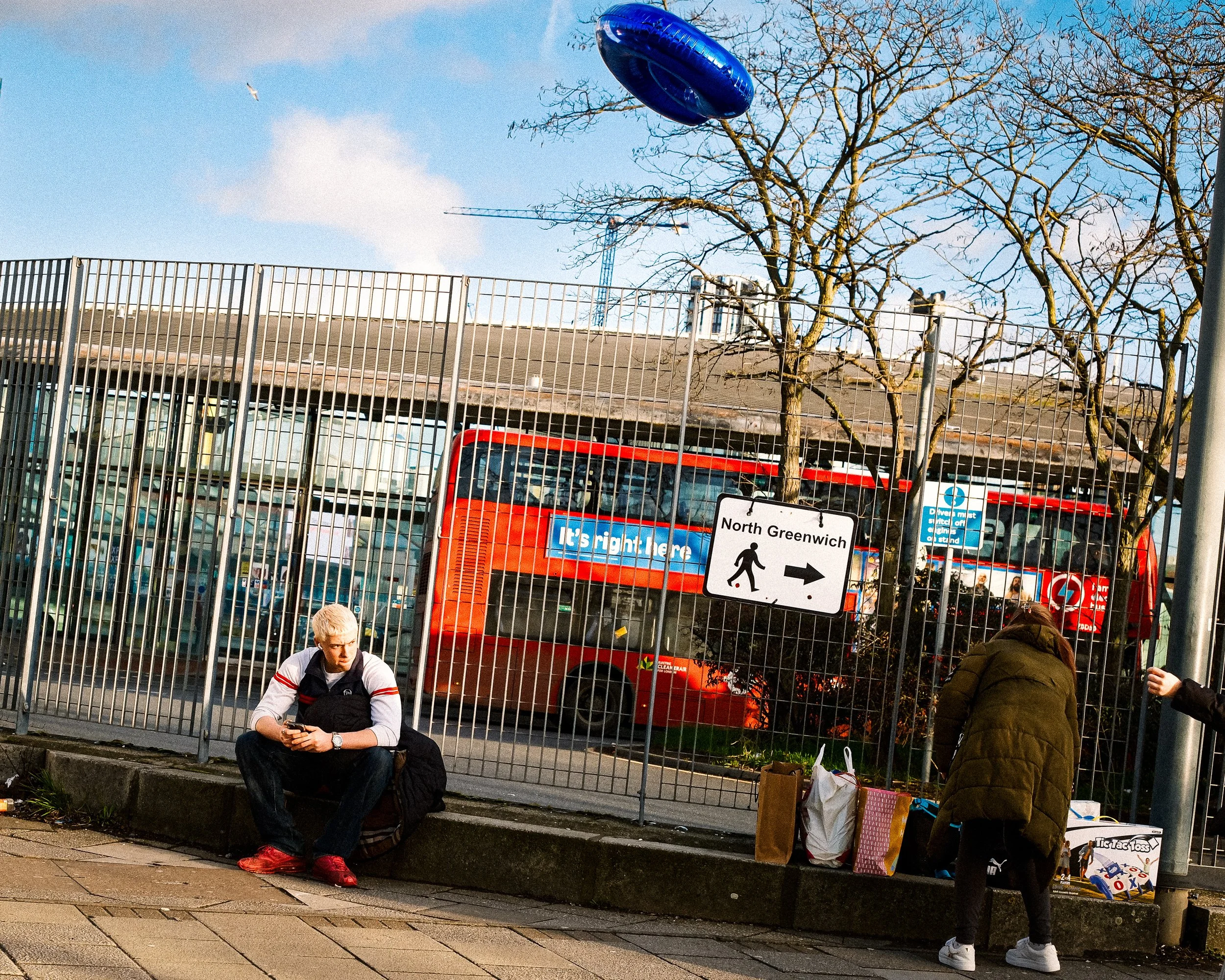 A person with blond hair sitting on a concrete ledge, looking at their phone, with bags around them. A woman with a brown coat is bending down near the bags. Behind the fence, there is a red double-decker bus and leafless trees, with a blue sky and a plane flying overhead.