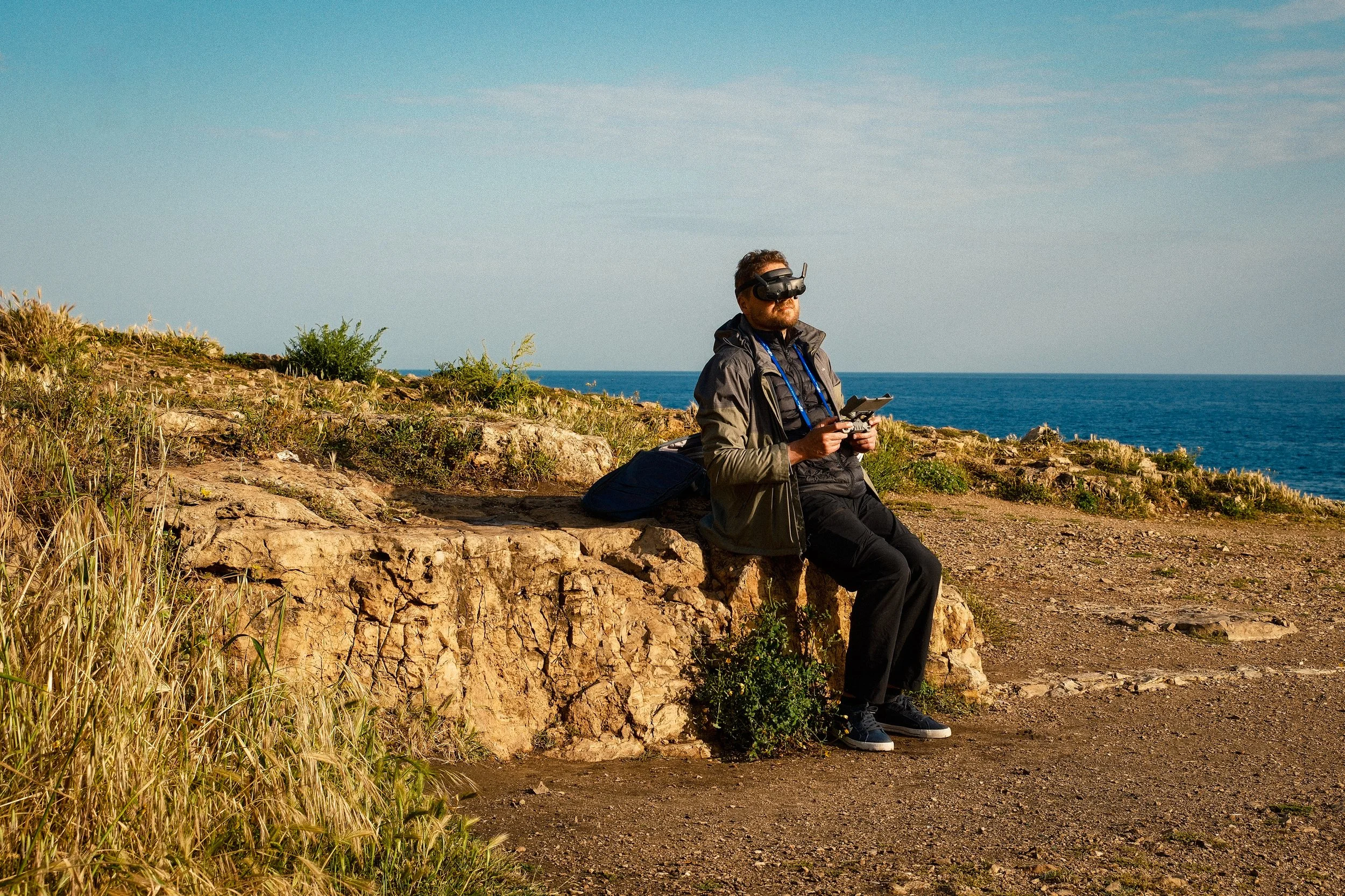 A man sitting on a rock ledge near the ocean, wearing a virtual reality headset and holding a remote controller, with a backpack beside him on the ground.
