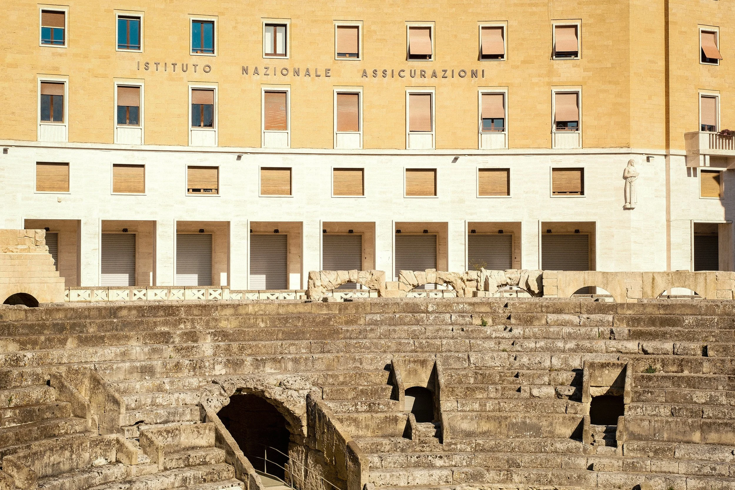 Exterior of a yellow building with multiple windows, some with shutters, and an inscription in Italian, 'ISTITUTO NAZIONALE ASSICURAZIONI.' In front, old stone steps and structures, possibly part of an ancient Roman theater or amphitheater.