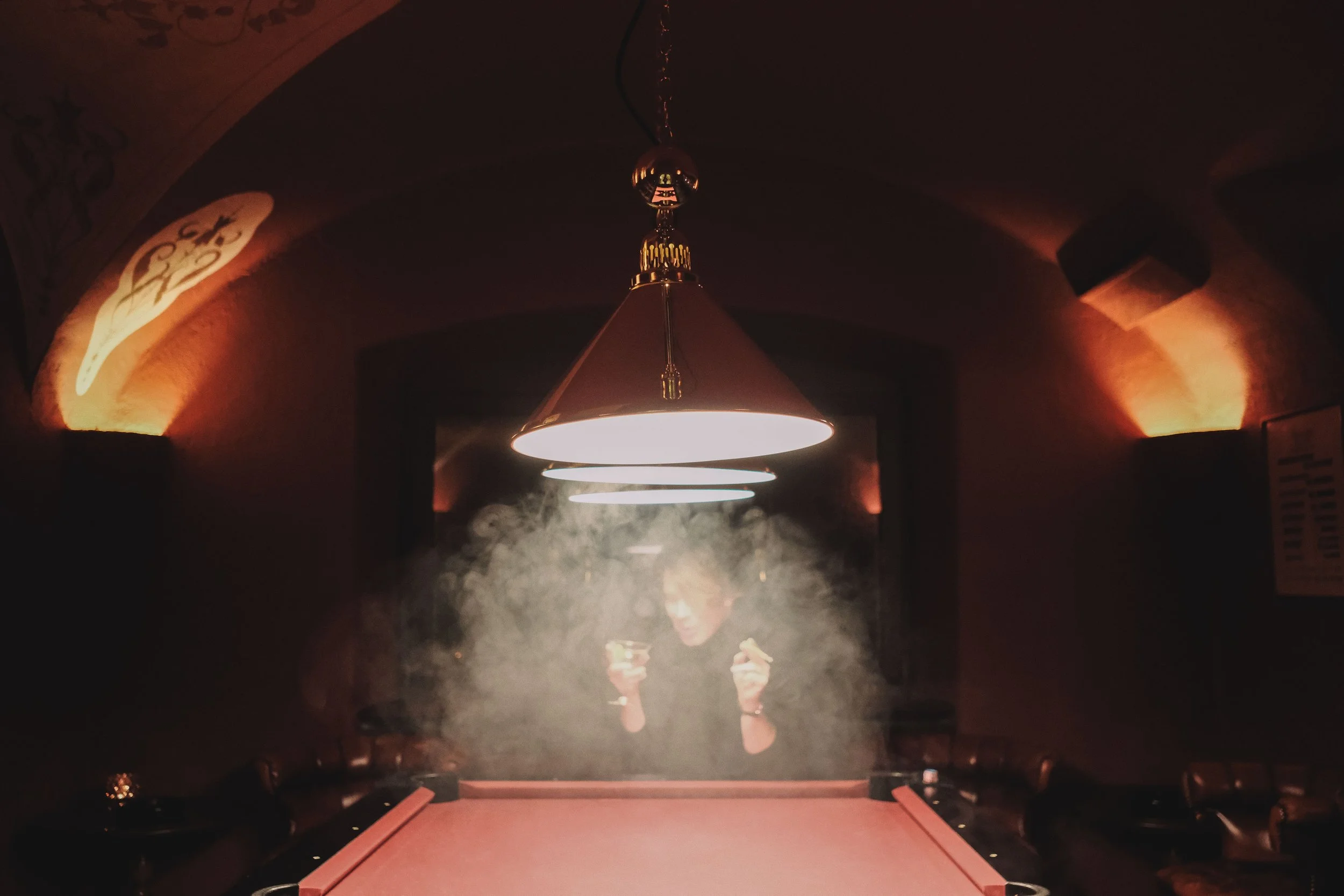 Dimly lit room with a red pool table, overhead lights, and a person holding drinks in the background surrounded by smoke.