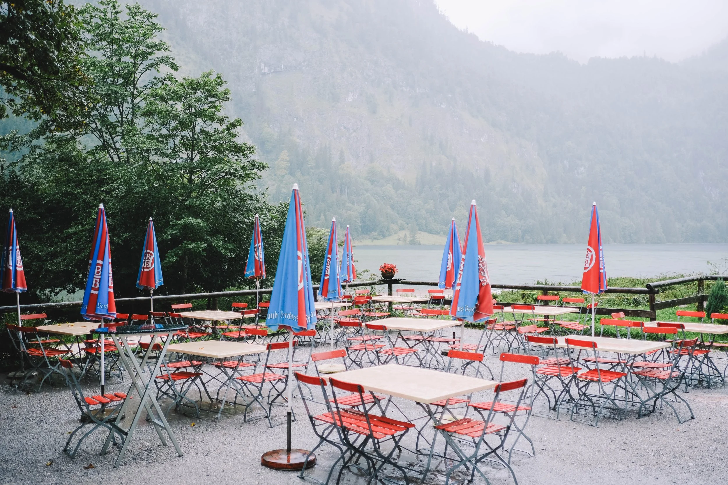 Outdoor seating area with red chairs and wooden tables, blue and red umbrellas, trees, and a misty lake view.