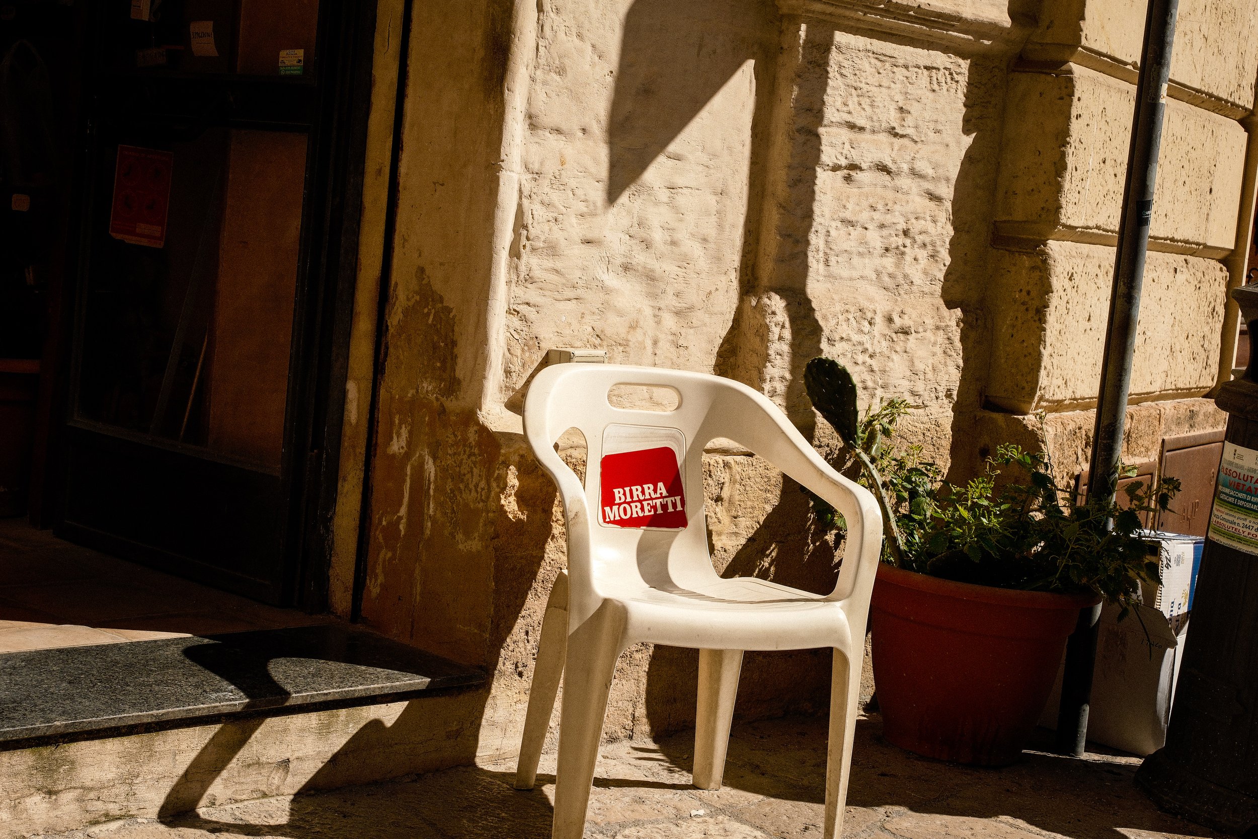 White plastic chair with a red sign that reads 'BIRRA MORETTI', positioned next to a large potted plant on a sidewalk in front of a stone wall and a shop window, with sunlight casting shadows.