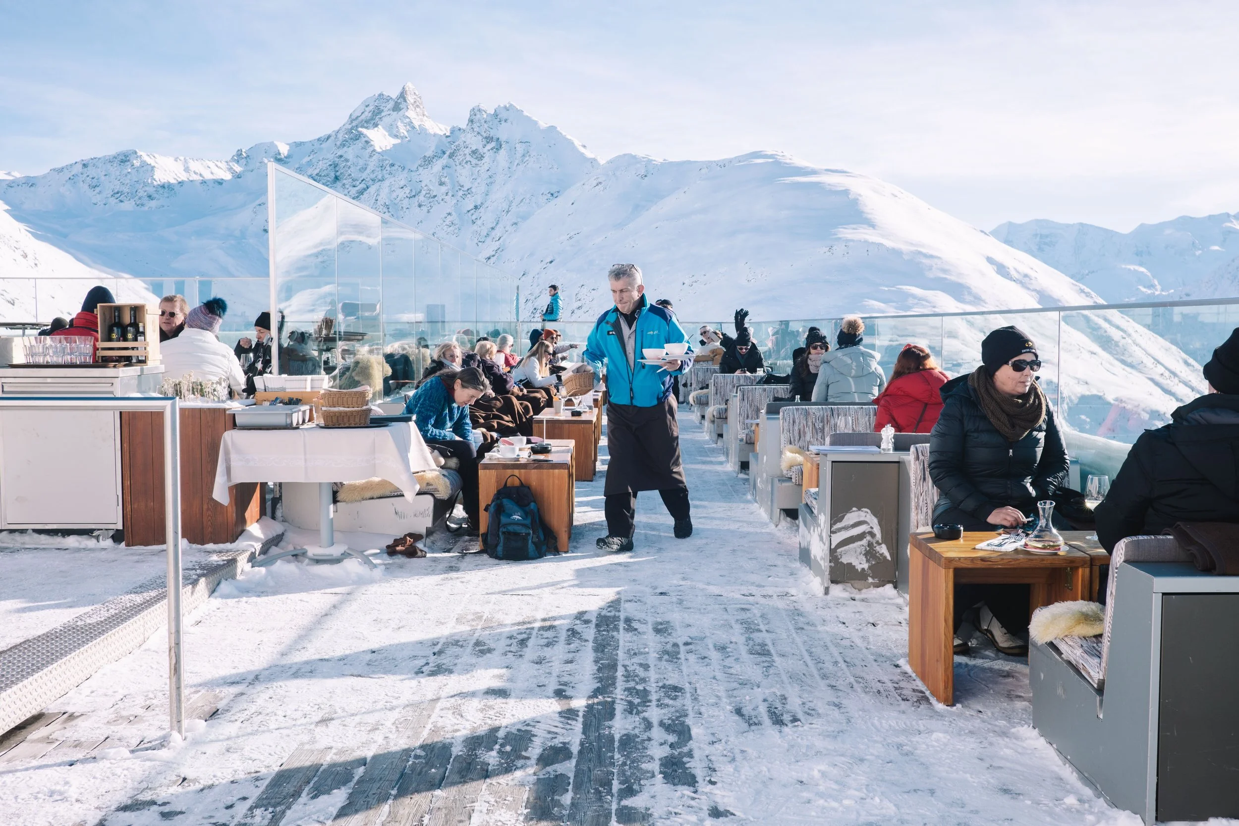 Outdoor cafe on a snowy mountain deck with people dining and enjoying the view, surrounded by snow-covered peaks. A waiter in a blue jacket serves customers.