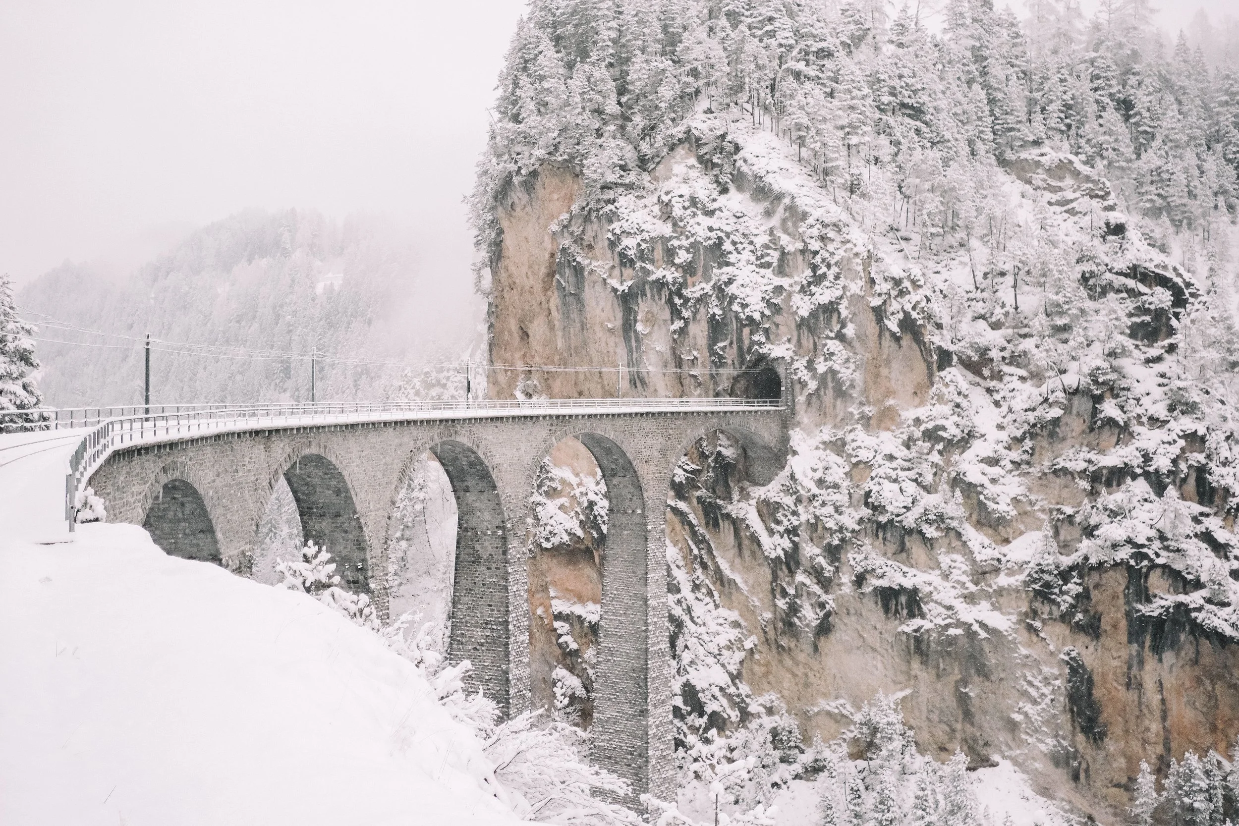 Snow-covered viaduct bridge with arches, surrounded by snowy cliffs and pine trees.