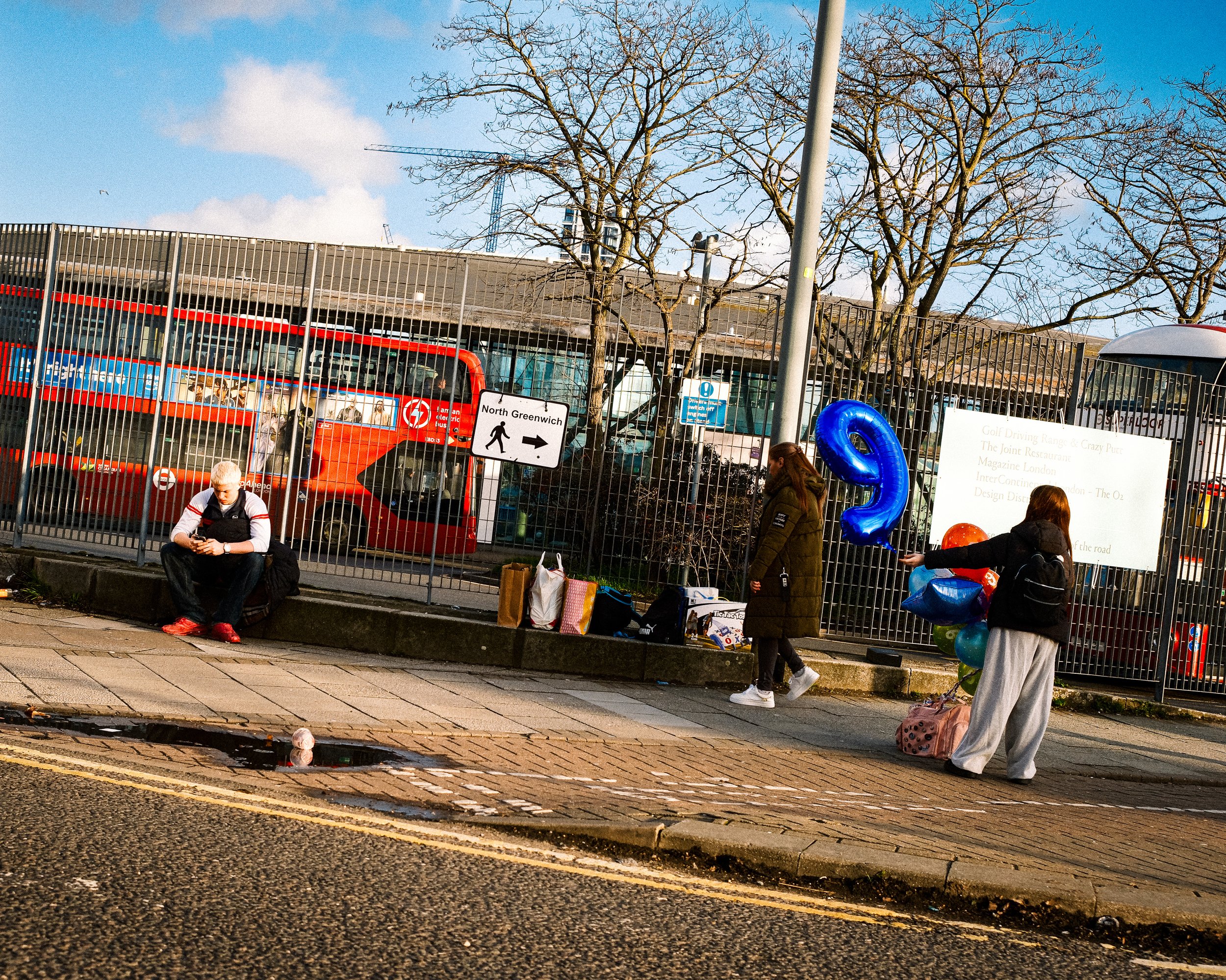 People waiting at a bus stop with motorcycles and a bus behind a fence. One person is sitting and looking at a phone, while two others hold balloons and bags. There is a large puddle reflecting a face mask on the street.