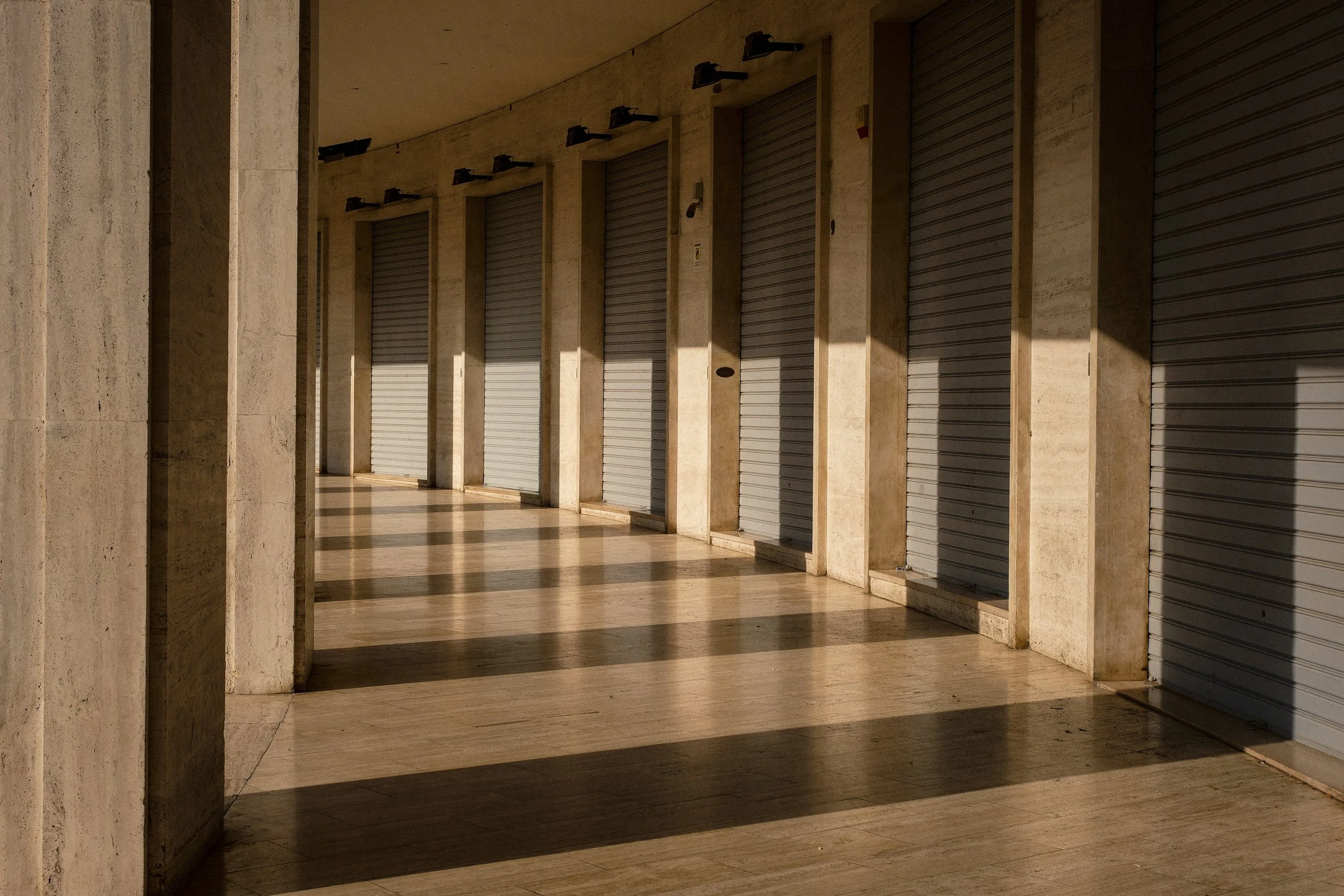 A row of closed metal shutters or gates in a corridor with marble walls and floors, illuminated by sunlight casting shadows.