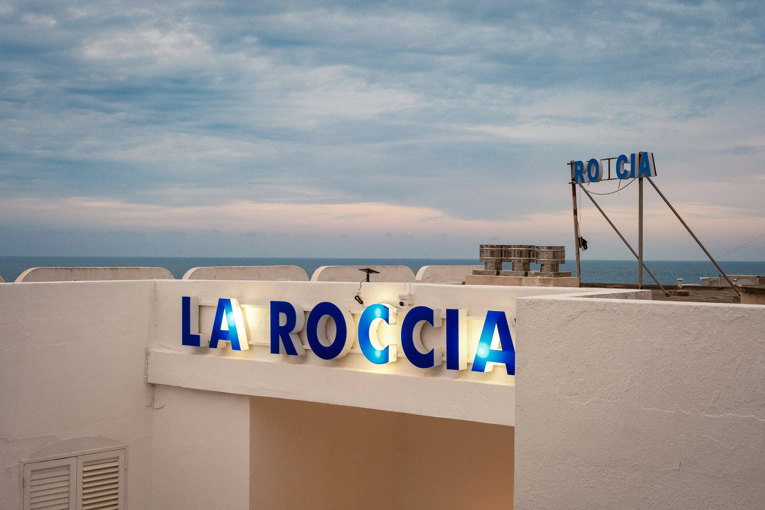 Sign that reads 'La Rocca' with a smaller sign above that says 'Trocia', overlooking the sea and a partly cloudy sky.