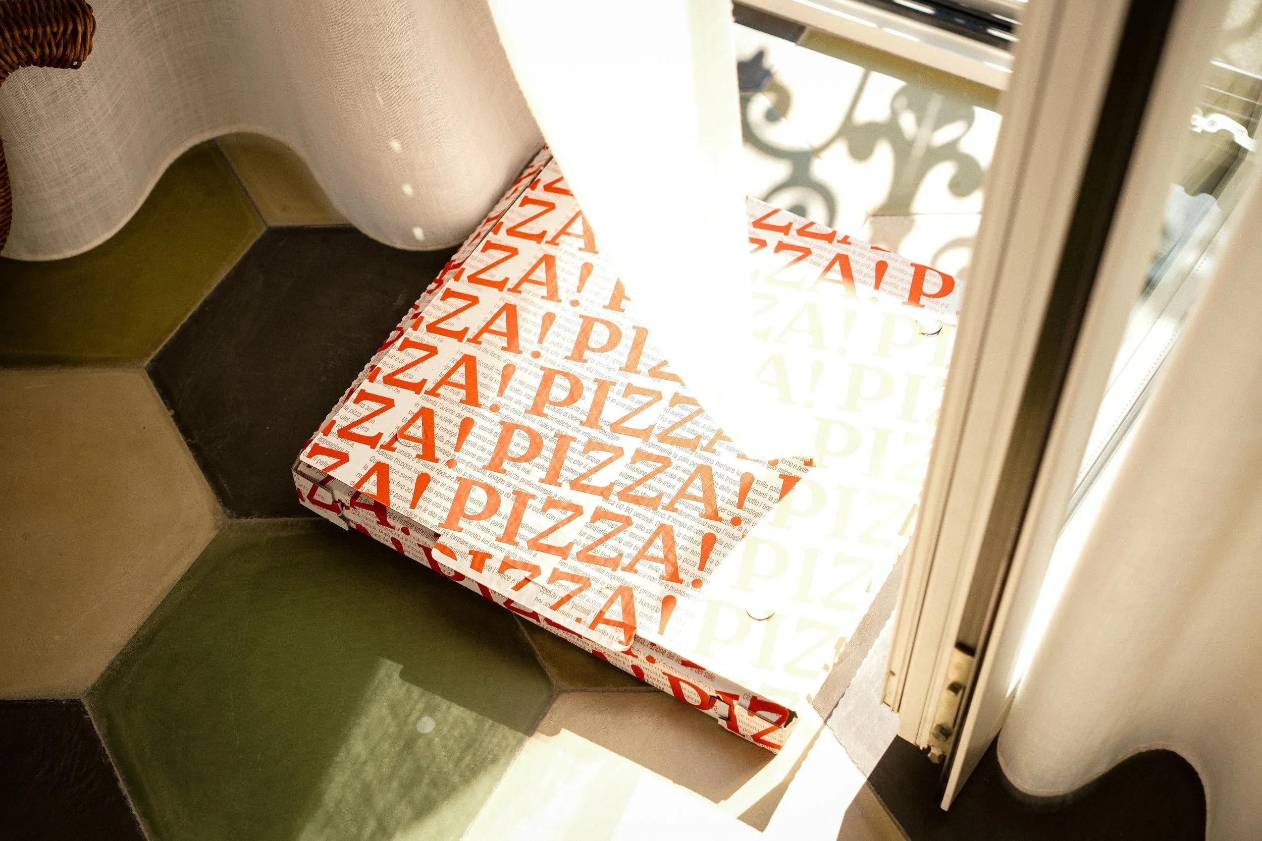 A pizza box with red and white lettering on a windowsill near a balcony door, with a curtain partially covering the window.