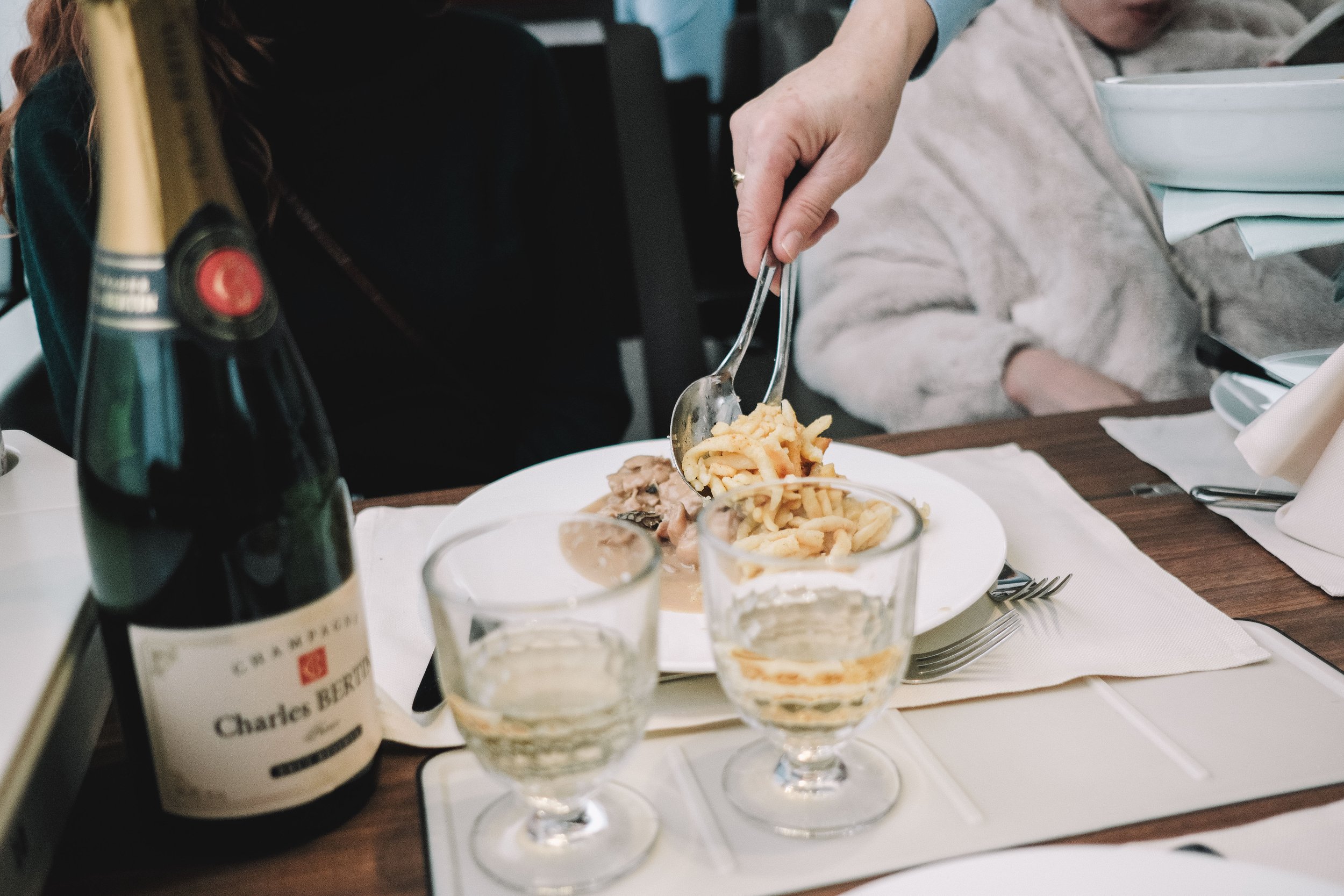A person serving fries with tongs onto a plate next to a bottle of champagne and two glasses of champagne on a table.
