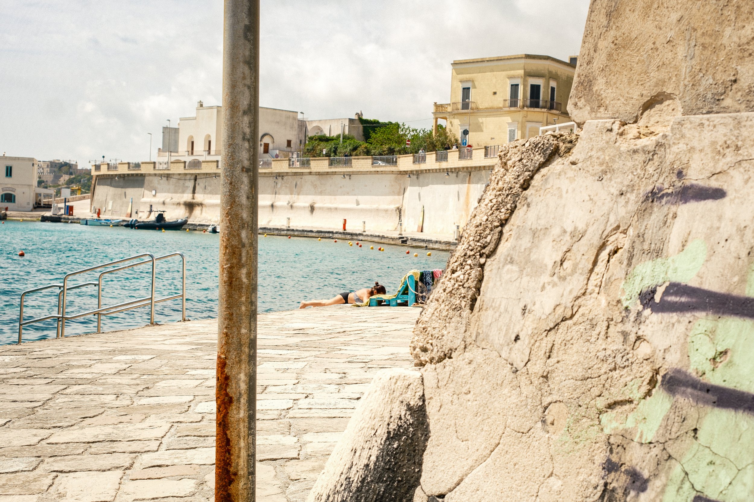 A woman sunbathing on a ledge near the water with colorful towels, a rusty pole, and a large stone wall in the foreground, with historic buildings and boats in the background.