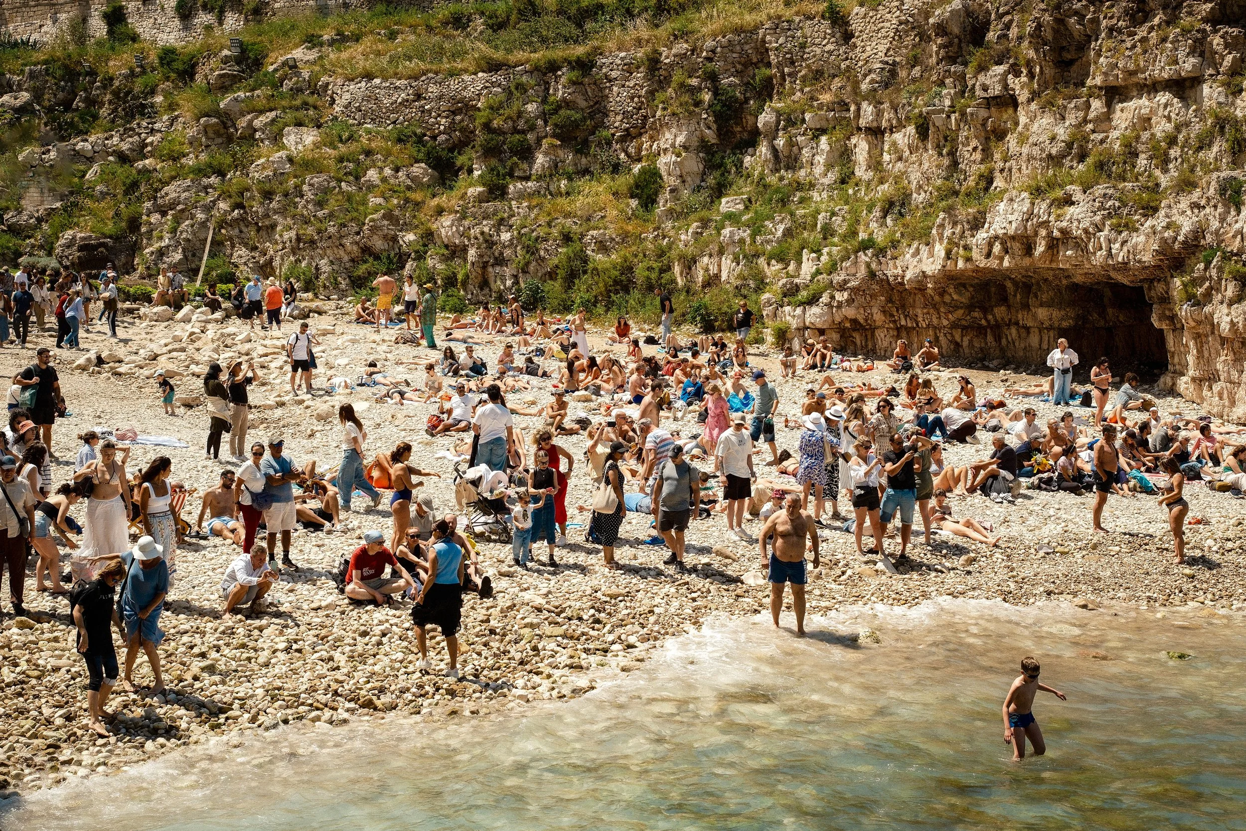 A crowded rocky beach with many people sunbathing, walking, and swimming near a cliffside.
