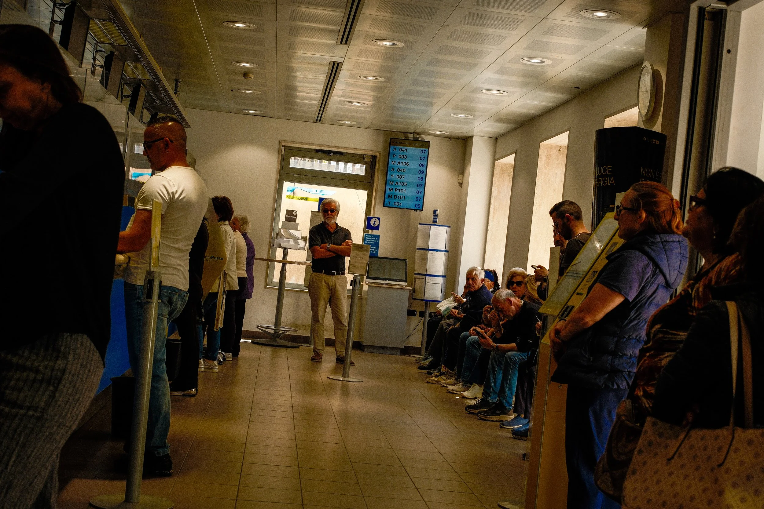 People waiting in line at a transportation or ticketing facility, with digital display boards showing schedules and numbers, and some individuals looking at their phones or talking.
