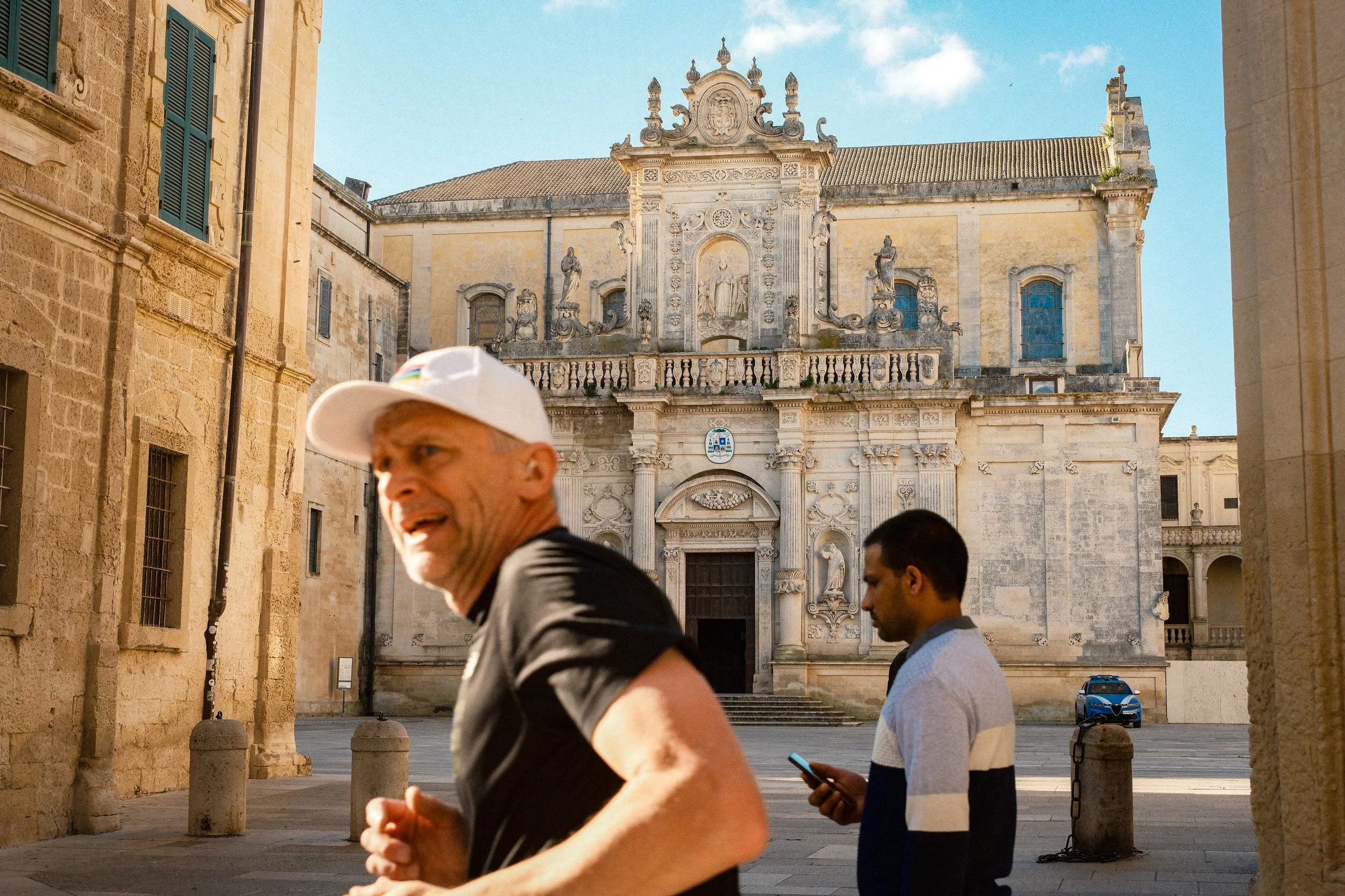 A historical building with ornate details and statues on its facade, viewed from a street where three men are walking; one in a black shirt and white cap, another in a striped shirt and glasses, and a third in a dark shirt.