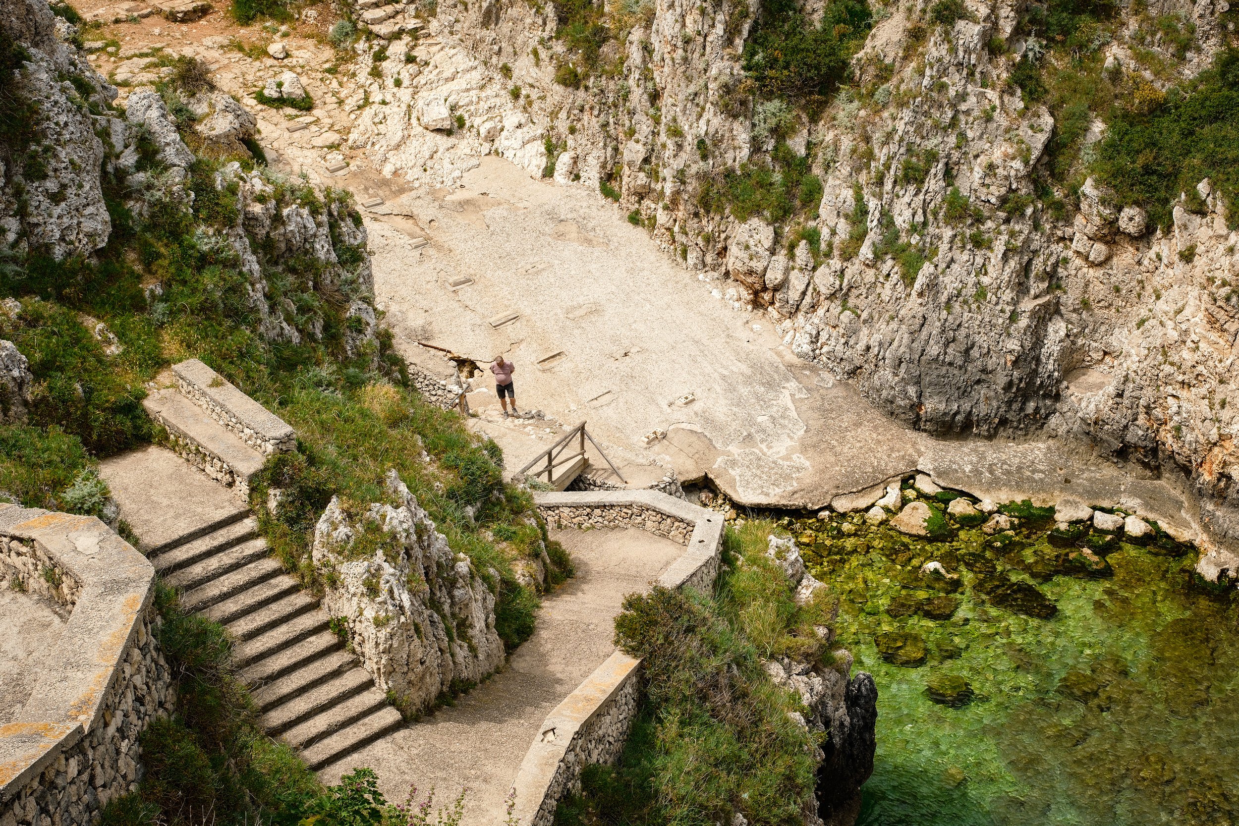 A person standing at the foot of a steep set of stone stairs leading up a rocky, grassy hillside in a narrow canyon with high rocky cliffs on each side.