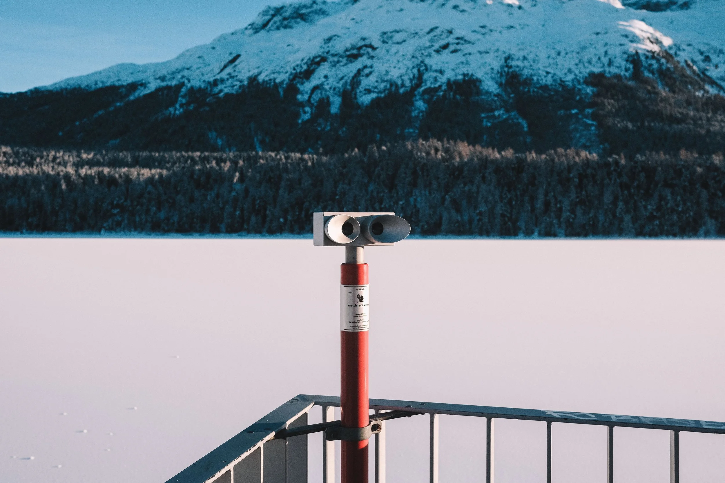 Binoculars on a red stand overlooking a snowy landscape with mountains and trees in the background.