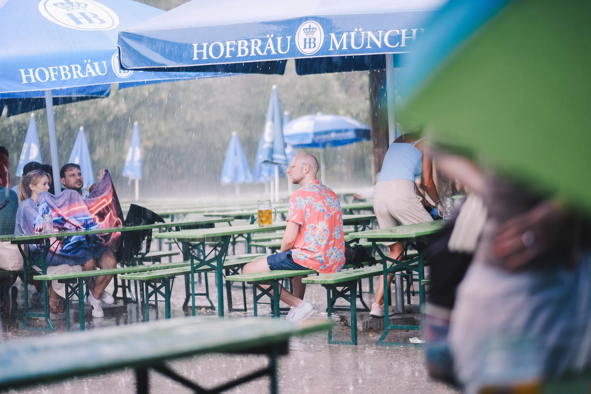 People sitting at outdoor tables under blue Hofbräu München umbrellas, experiencing rain. One person is wrapped in a colorful blanket, while another in a floral shirt sits nearby, with blurred movement in the foreground.