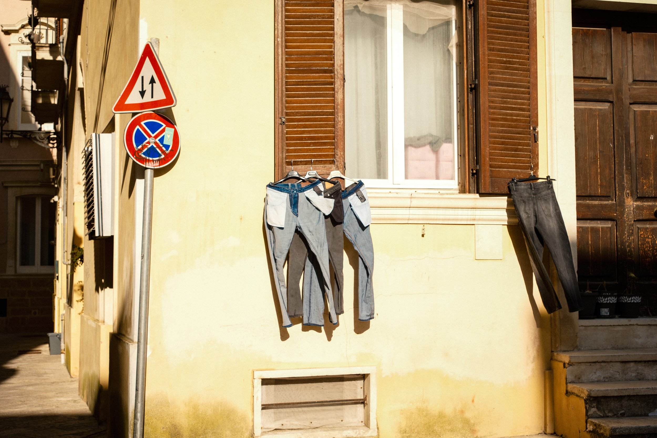 A yellow building with a window and open wooden shutters. Clothes, including pants and jeans, are hanging on a line outside. There are traffic and parking signs next to the building.
