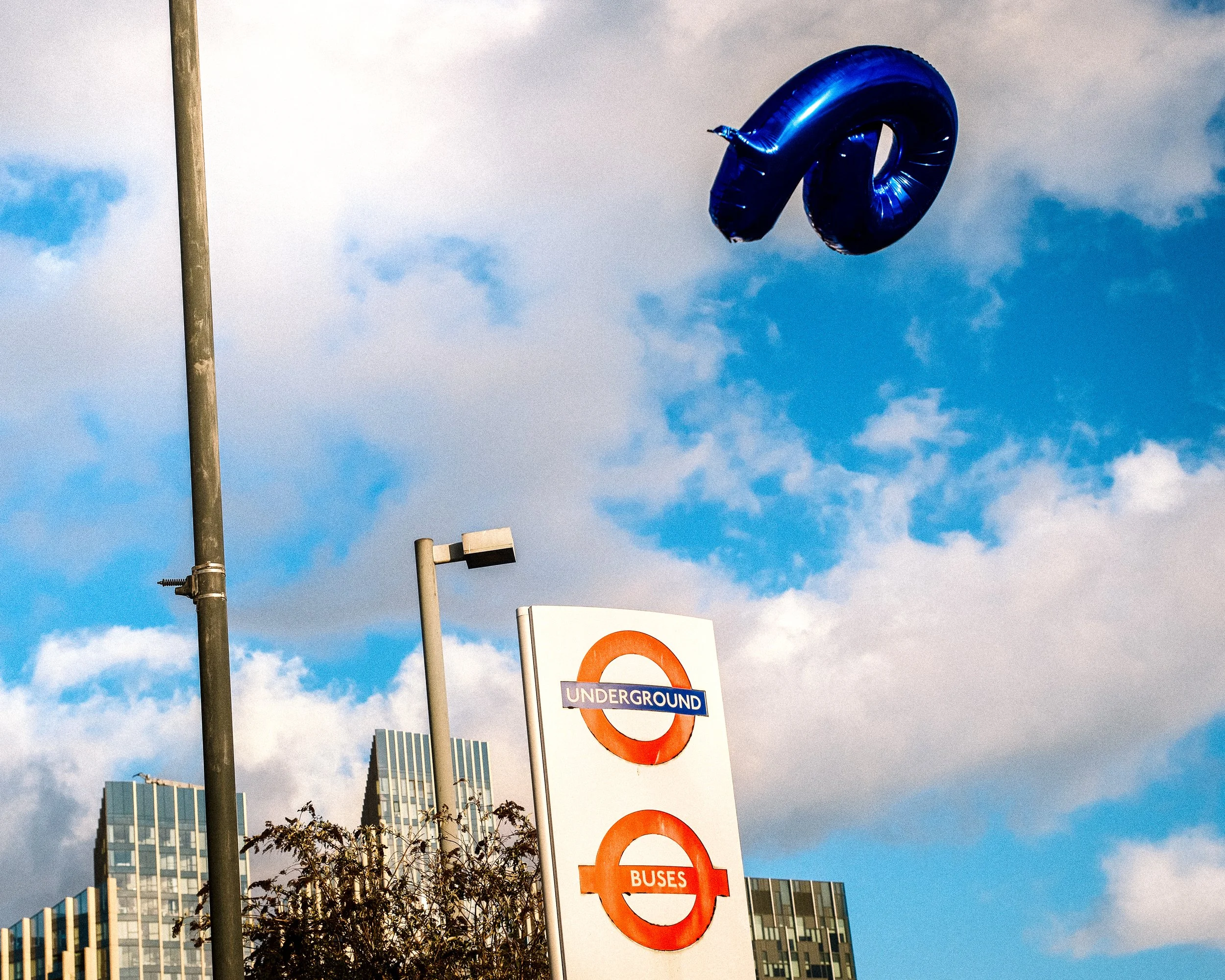 A large blue number 0 balloon floating in the sky near an underground subway sign, with modern buildings and a cloudy sky in the background.