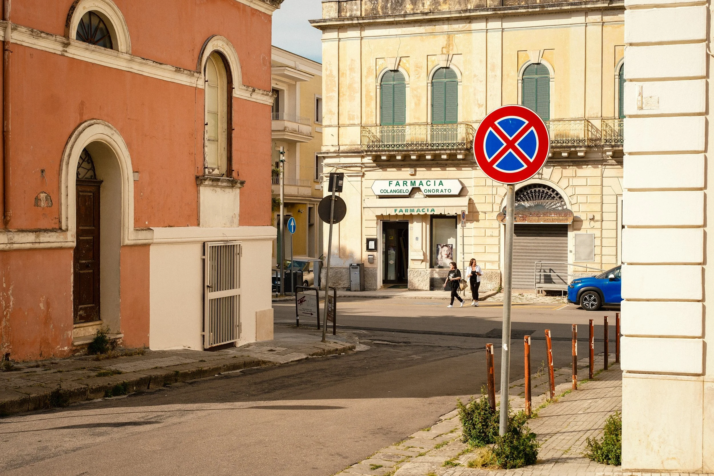 Street scene with pastel-colored buildings, a no parking sign, a pharmacy, and people walking on the sidewalk.