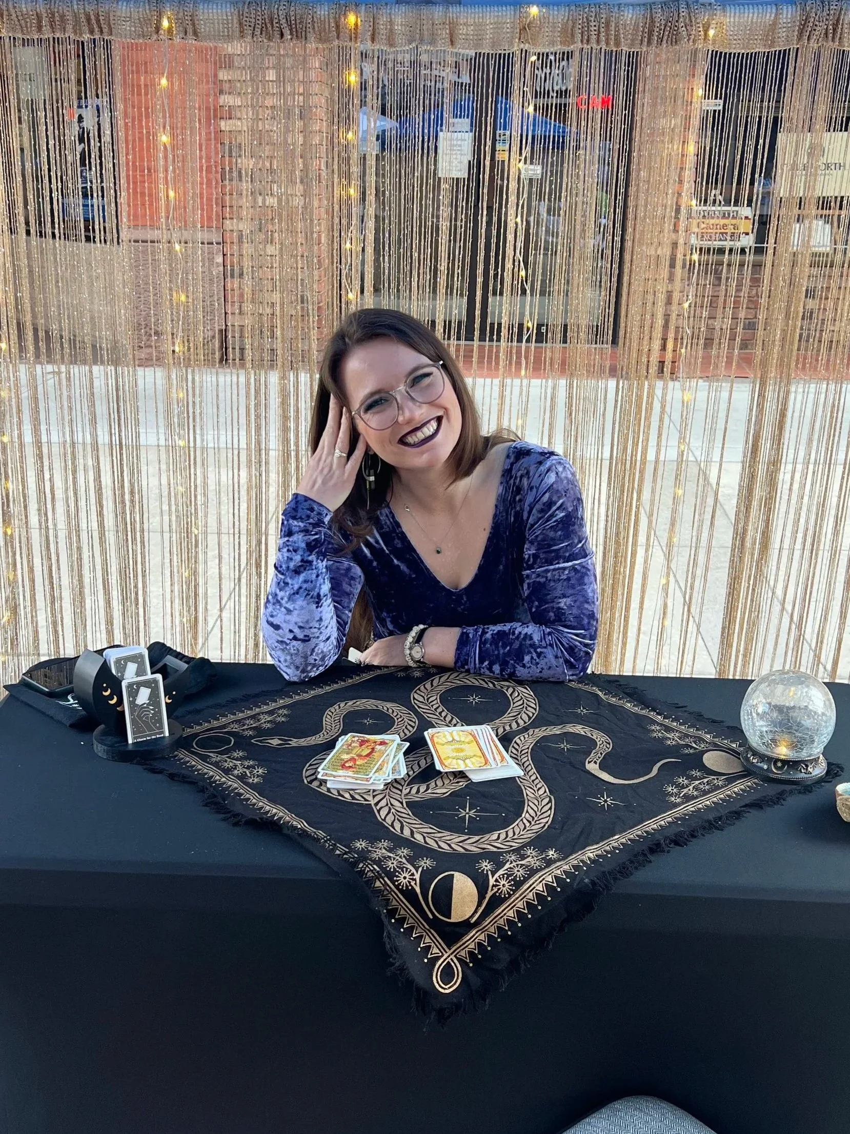 Woman with glasses smiling at a tarot reading table with tarot cards, crystal ball, and mystical items, in front of a beaded curtain with fairy lights.