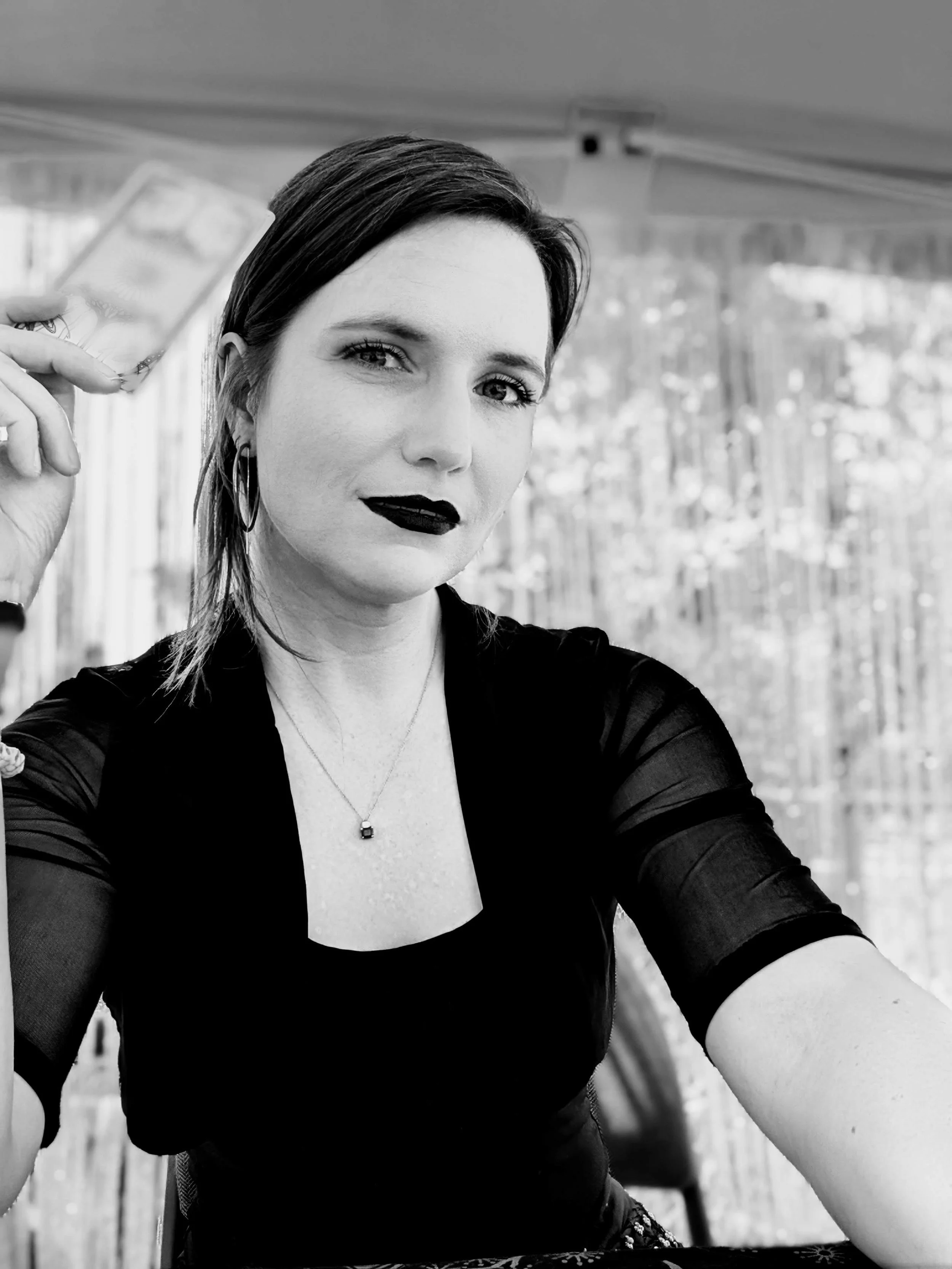 A woman with dark hair pulled back from her face, holding a tarot card in her right hand, wearing black lipstick, hoop earrings, a necklace with a small pendant, and a black outfit with sheer sleeves, in front of a background of shiny streamers.