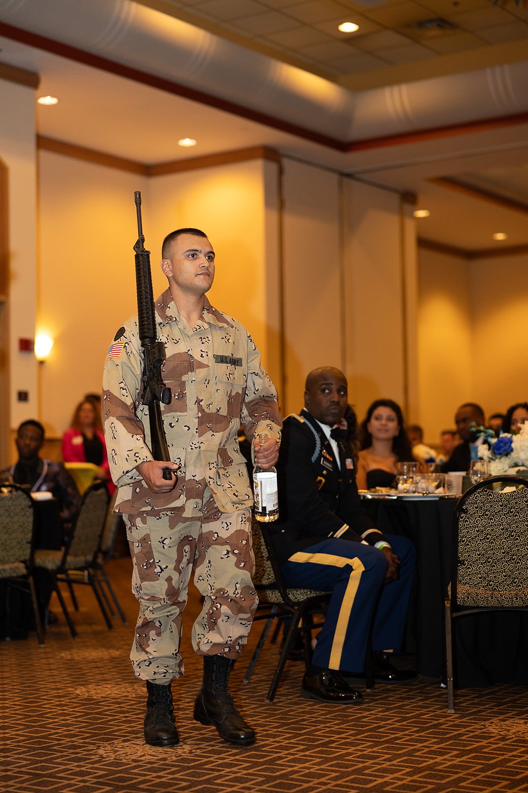A soldier in camouflage uniform holding a firearm walks through a formal event with seated attendees in military dress and civilian attire in the background.