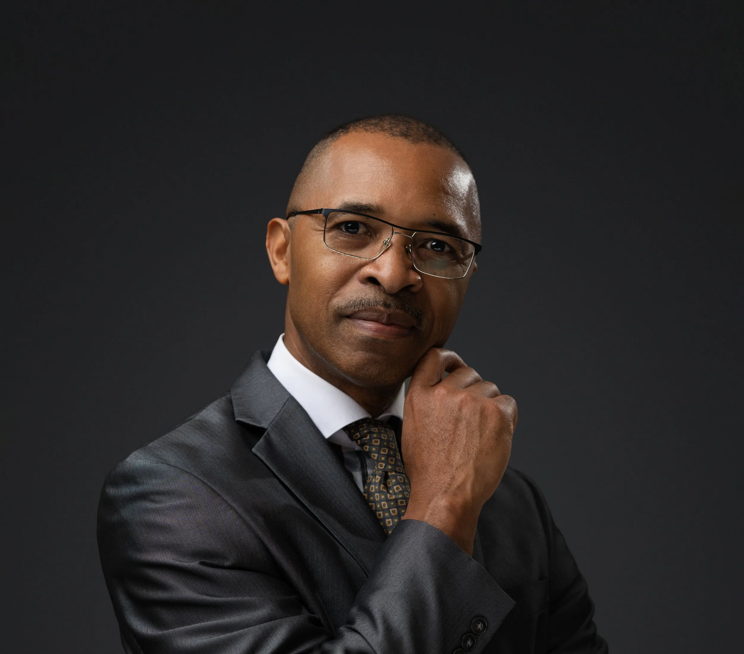Mid-adulthood man in a suit and glasses, posing thoughtfully against a dark background.