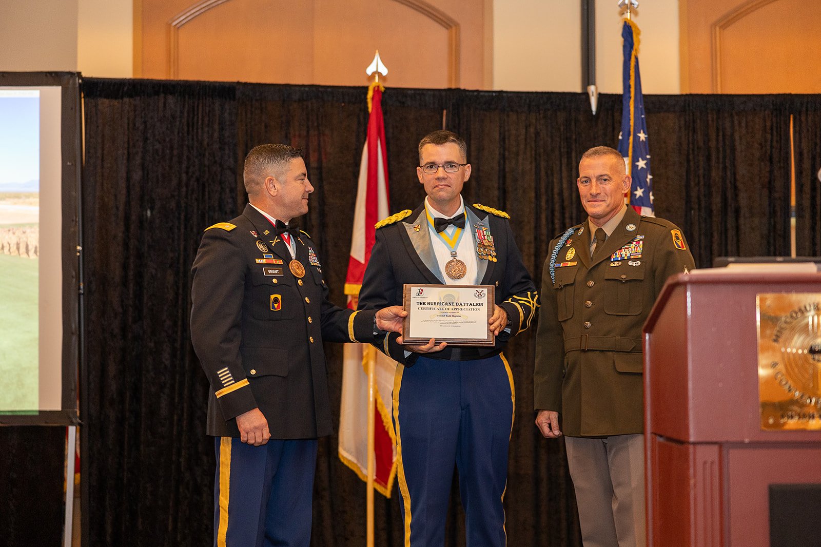 Three military officers standing on stage during an award ceremony, with one holding a certificate and medals. The background features black curtains and American and military flags.