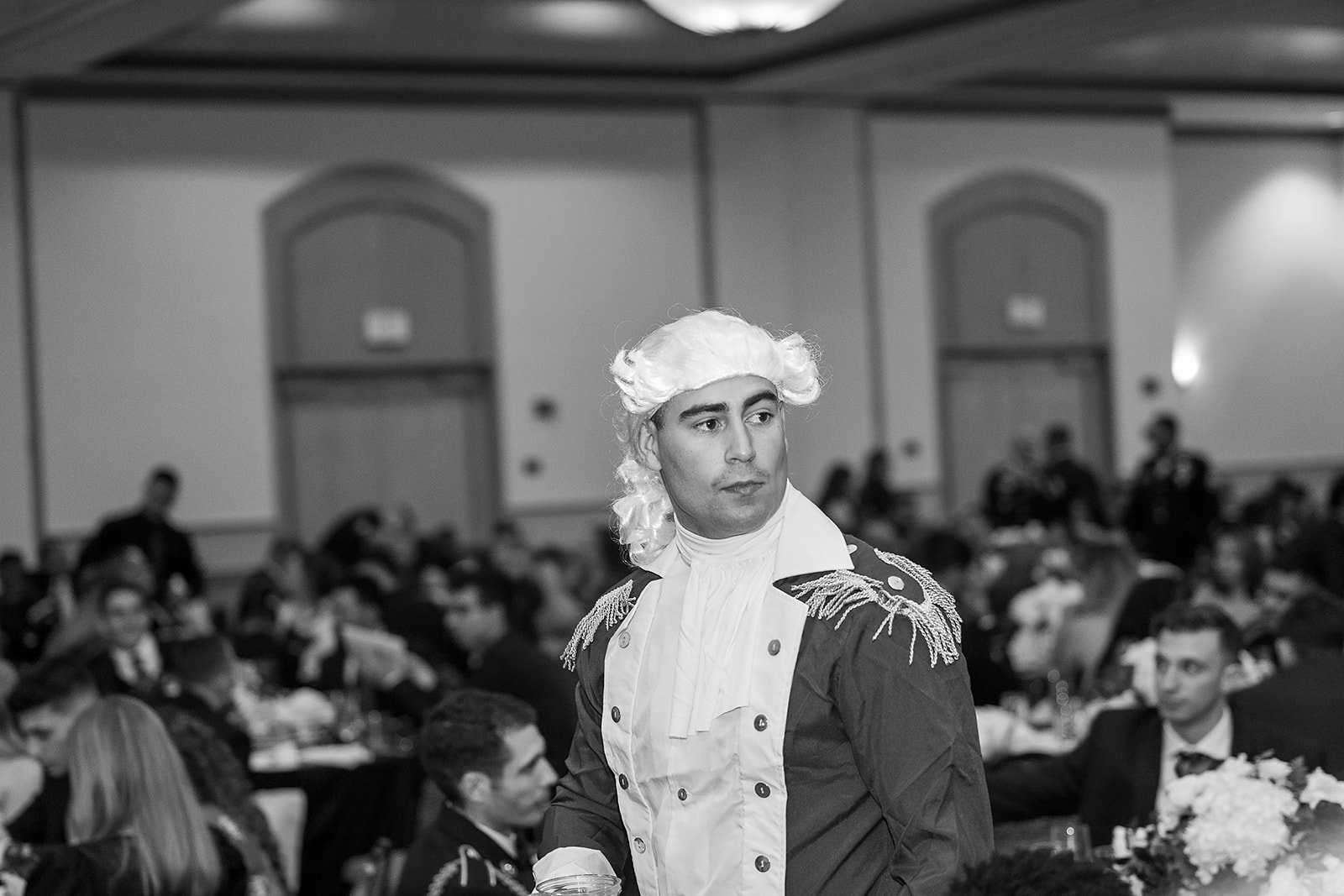 A man dressed in 18th-century historical costume, with a powdered wig, standing in a banquet hall filled with seated guests dressed in formal attire.