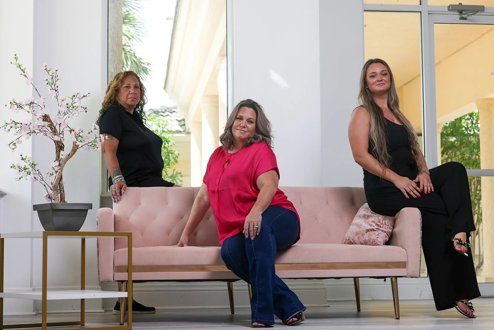 Three women with medium to light skin tones sitting and standing around a pink sofa in a bright indoor space with large windows and a potted cherry blossom tree.