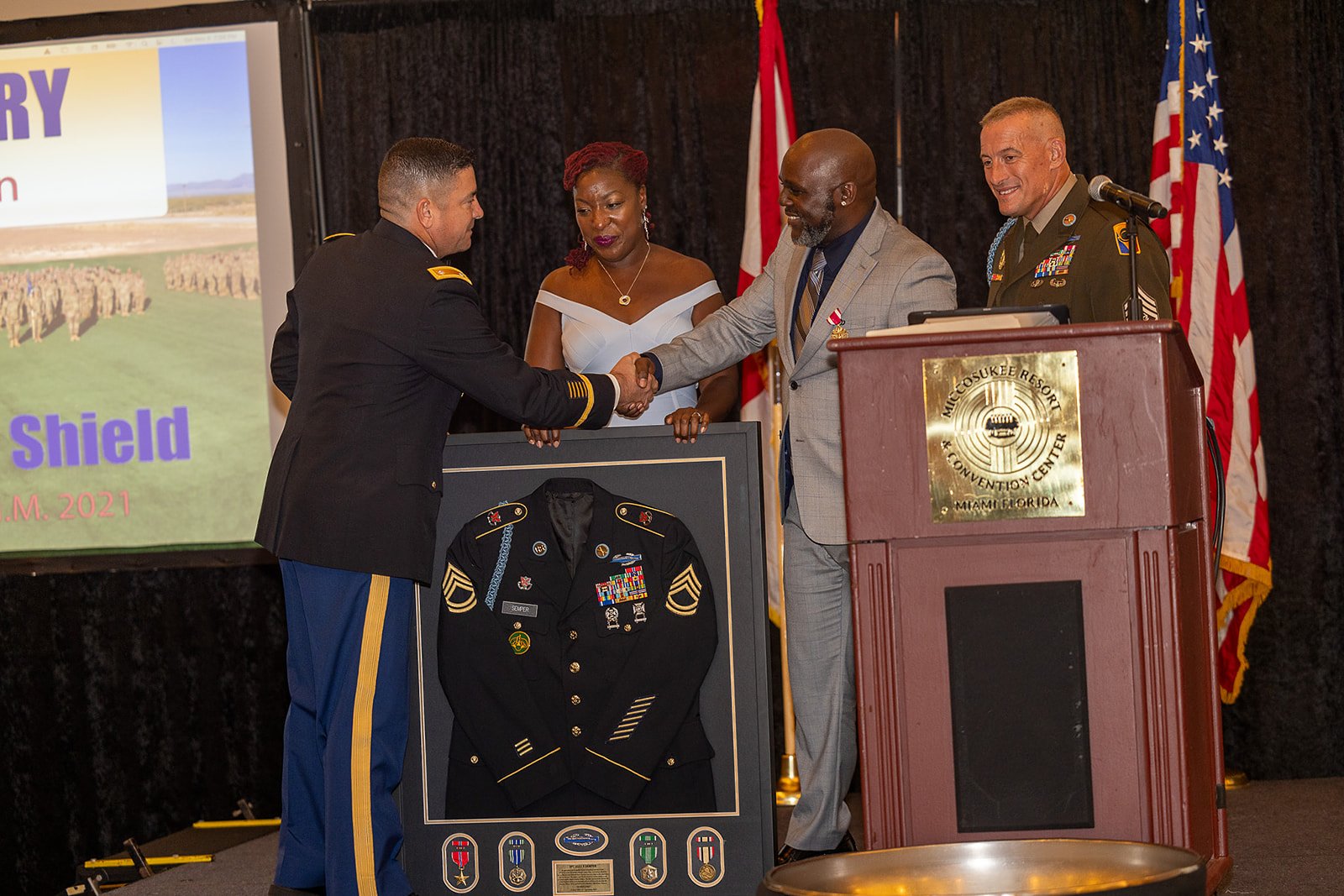A formal ceremony at the Micosukee Resort and Conference Center in Miami, Florida, where a man in military uniform is shaking hands with another man in a gray suit. A framed military uniform with ribbons and badges is displayed in front of them. Ther
