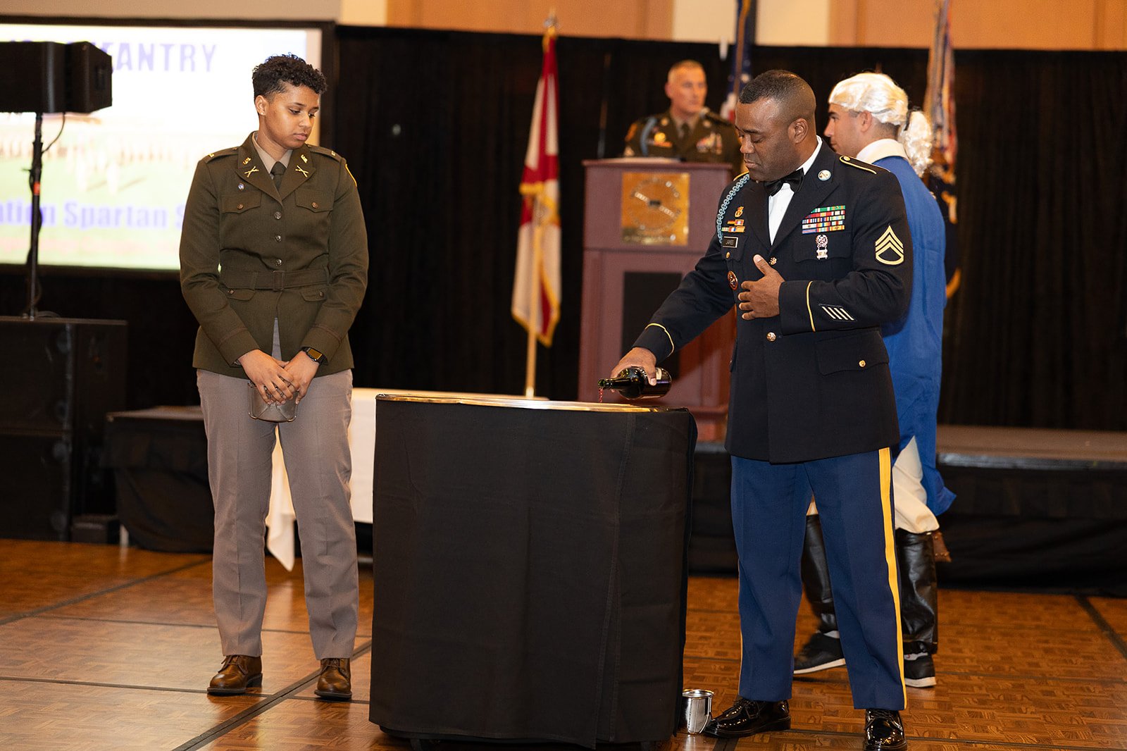 A military award ceremony with a man in a veteran uniform pouring wine and a young woman in uniform standing nearby, with a stage, flags, and a screen in the background.