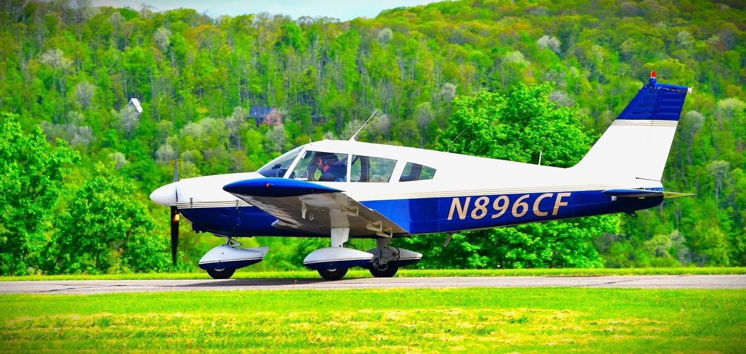 A small white and blue aircraft on a runway with green trees and hills in the background.