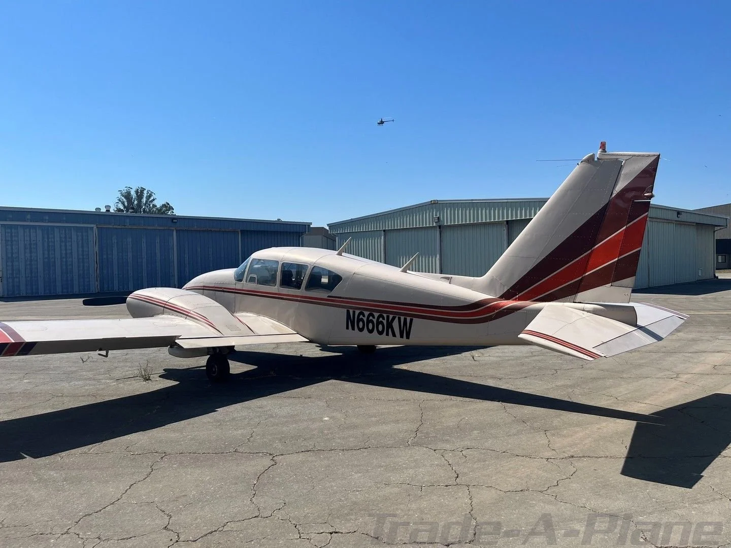 Small white private aircraft with red and black stripes parked on a tarmac in front of hangars, with a helicopter flying in the blue sky in the background.