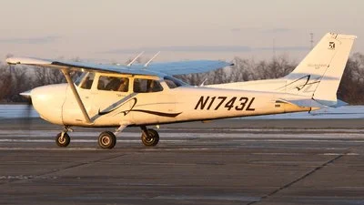 Small aircraft with tail number N1743L parked on runway with trees and cloudy sky in background.