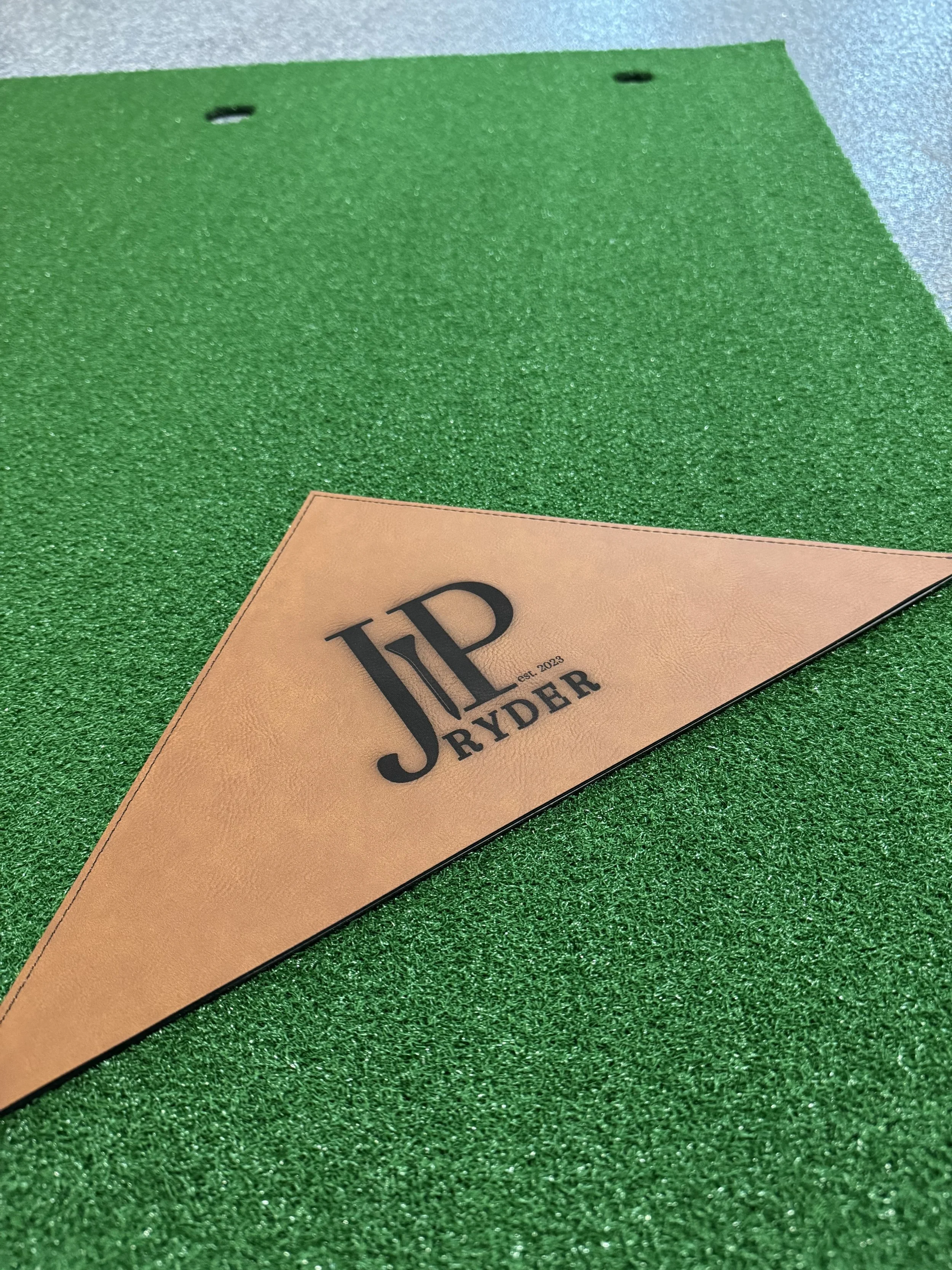 Close-up of a golf tee with a leather label that reads "J Ryder" on a textured green surface.